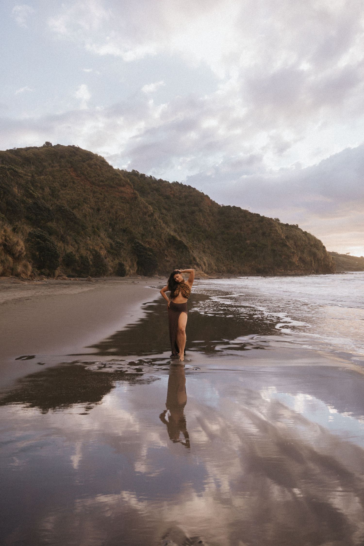 A silhouette walks along a reflective beach at sunset with dramatic cliffs and cloudy skies in the background.