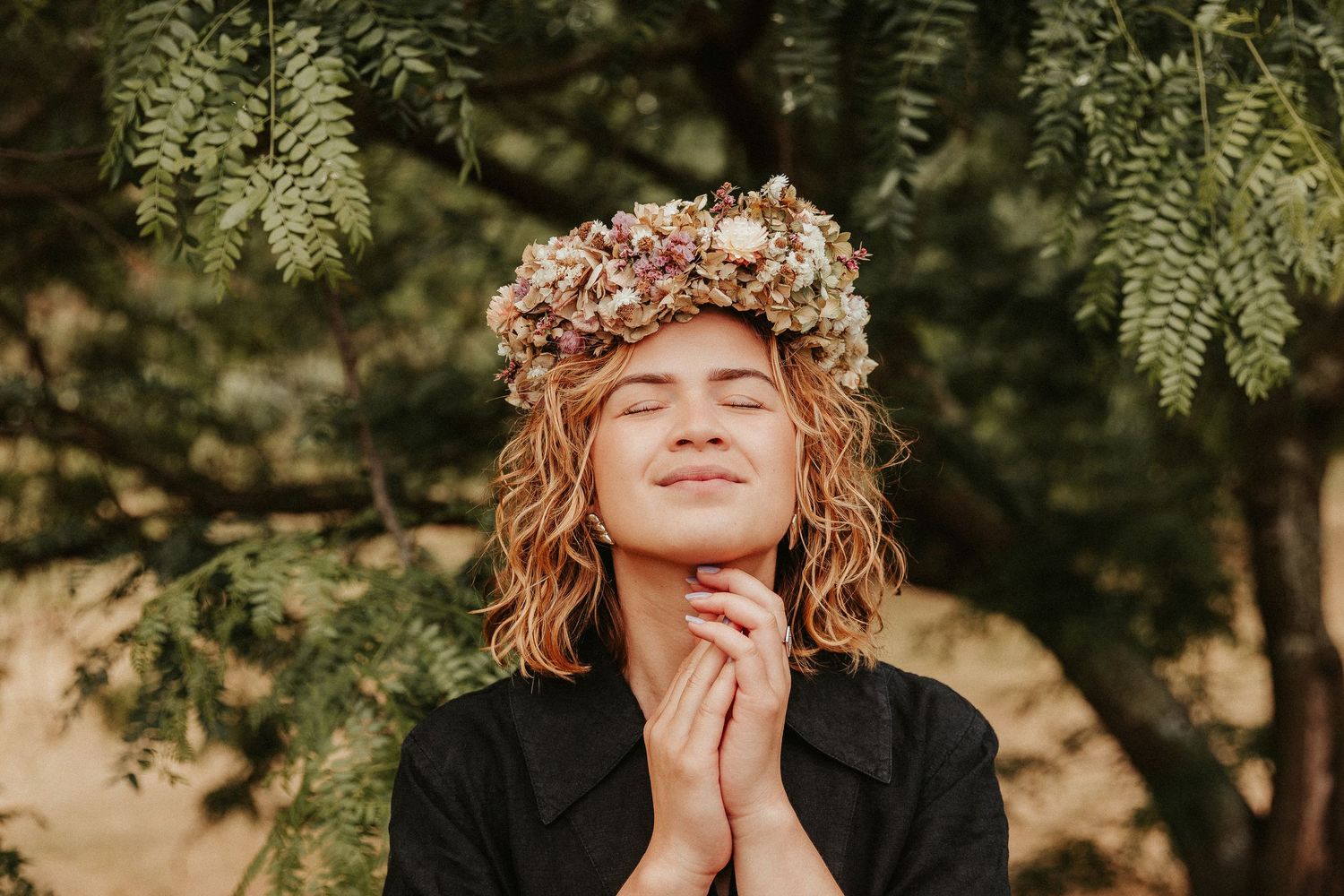 Person wearing floral crown poses peacefully outdoors among green foliage.