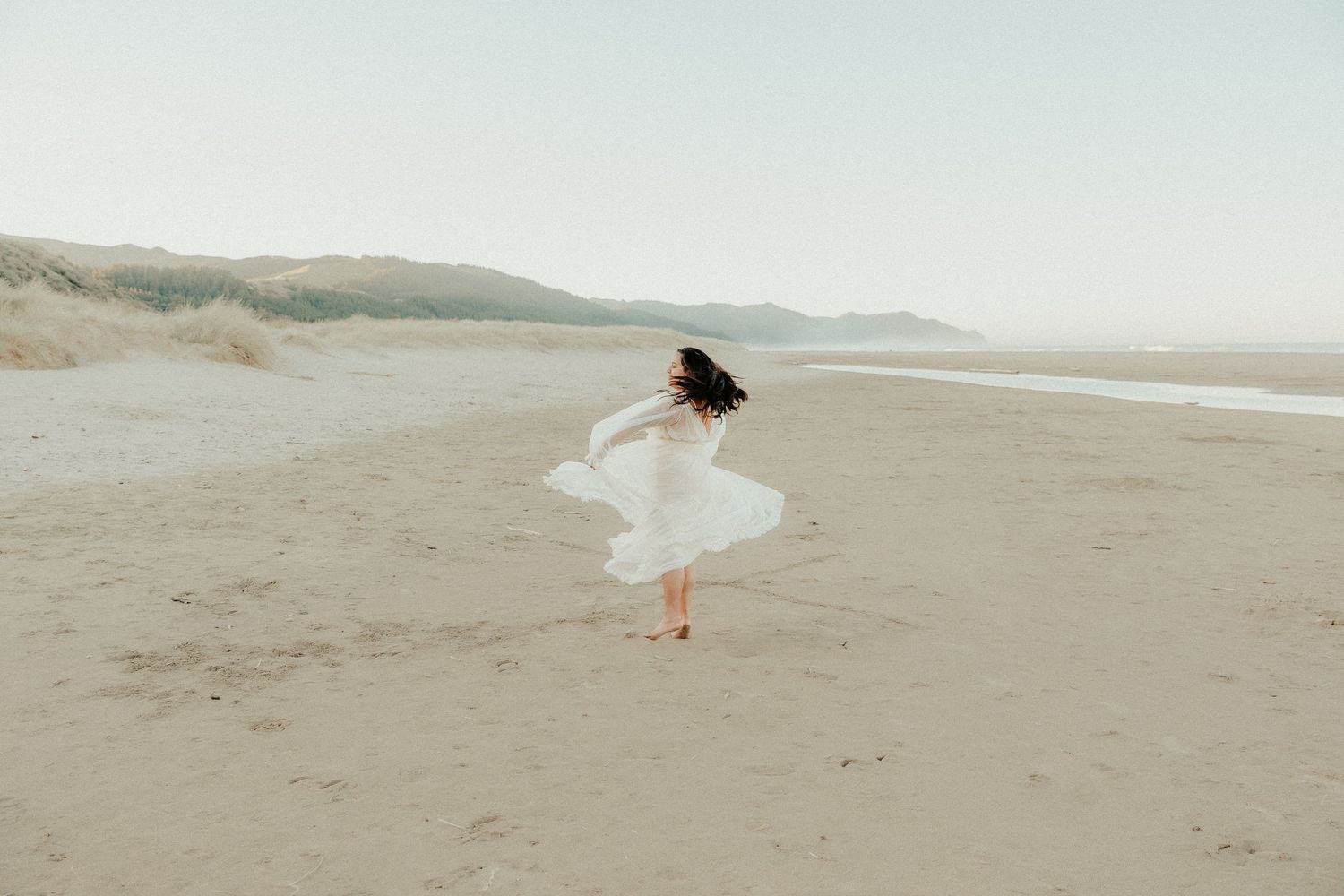 Figure in white dress dances freely on empty beach with mountains in background.