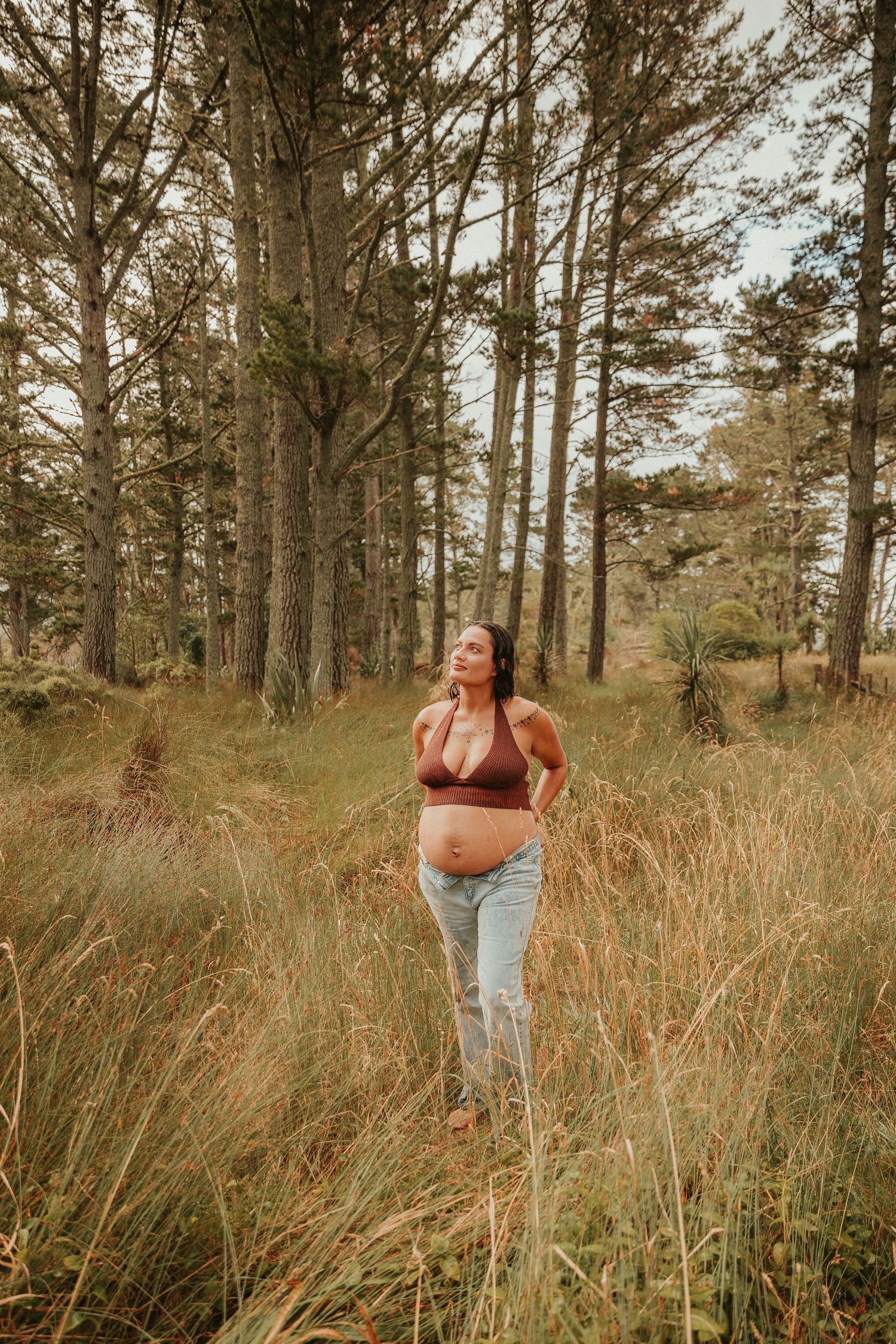 Pregnant woman in casual attire poses in tall grass with pine trees in the background during golden hour.