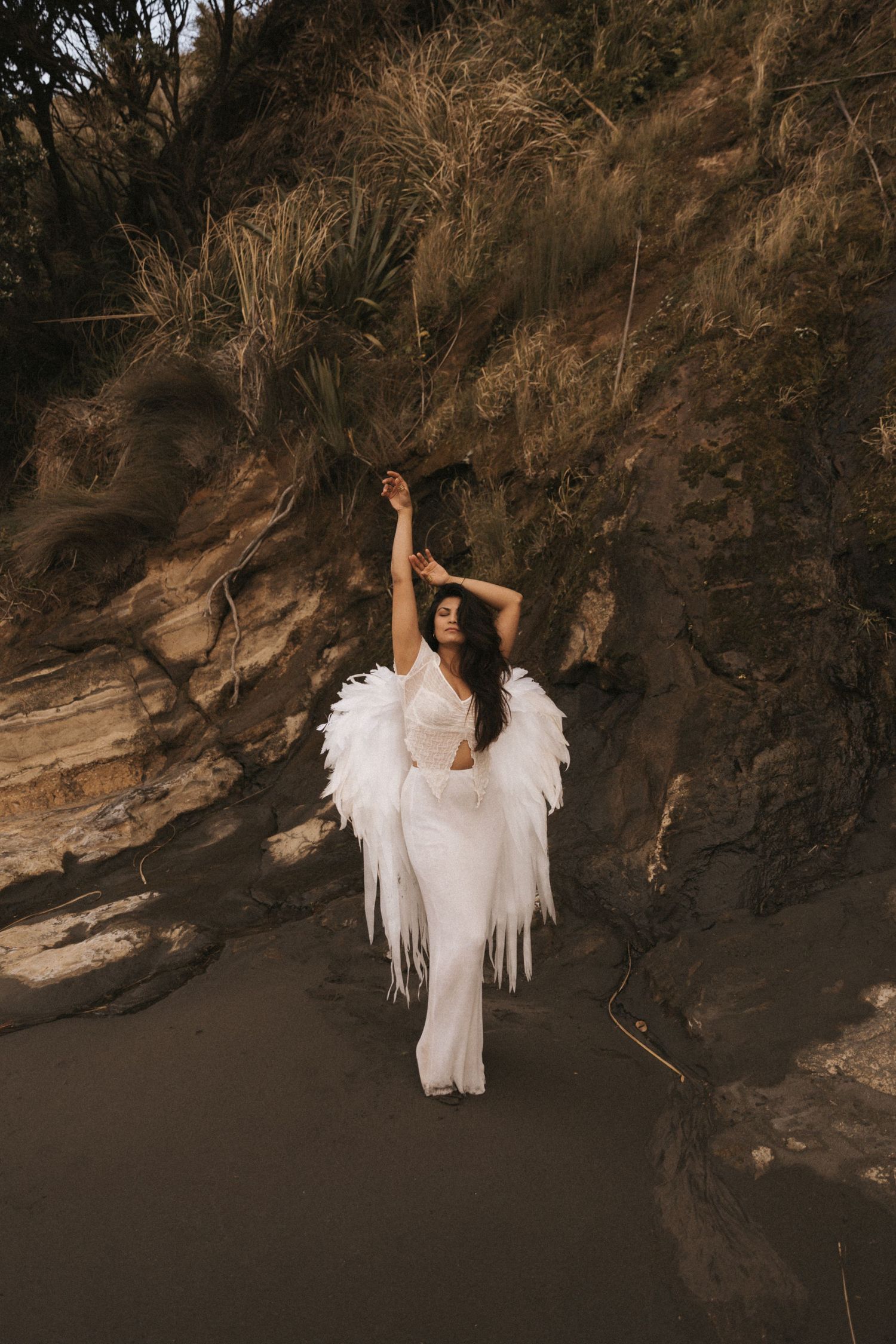 A figure in a flowing white dress and angel wings poses dramatically against a dark rocky cliff at sunset.