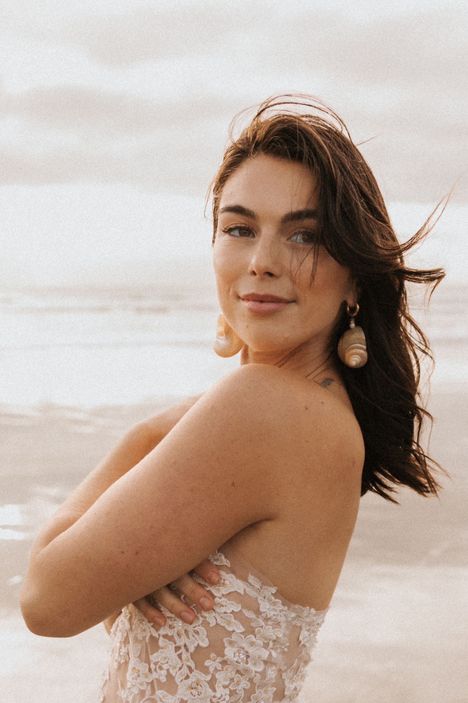 Woman in white lace dress poses gracefully on a beach during sunset.