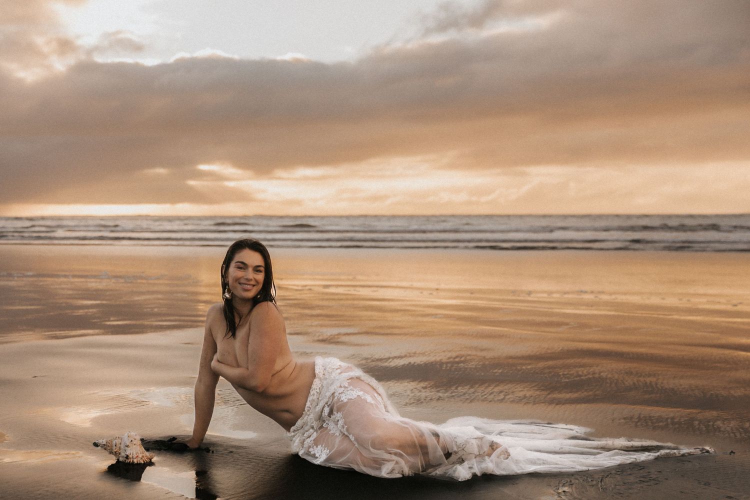 A white wedding dress flows across dark sand as waves crash on a moody beach at sunset.