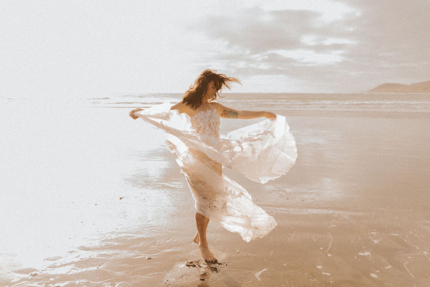 Woman dances freely in white lace dress on misty beach.