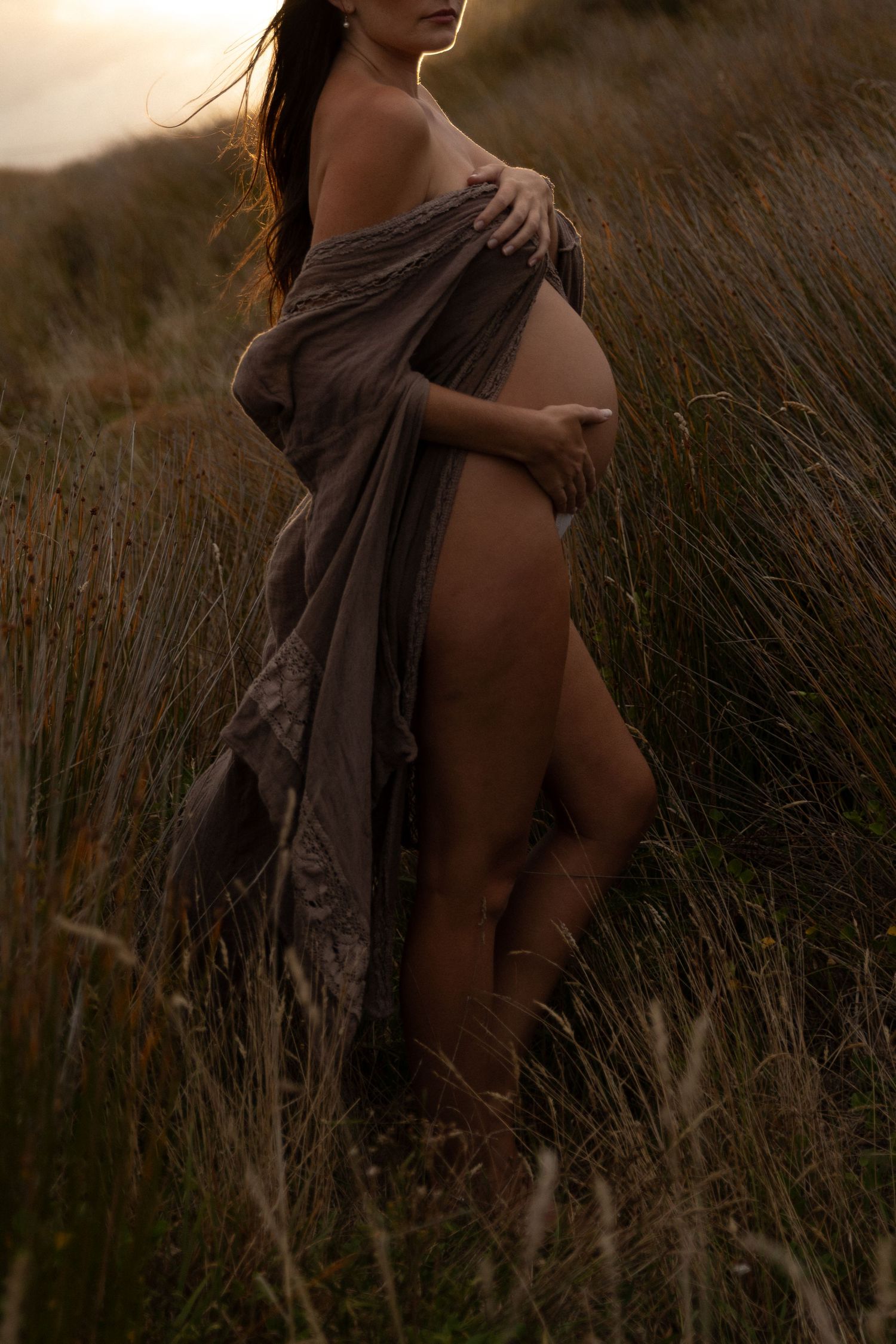 Artistic maternity session with dark fabric in golden hour lighting among tall grasses.