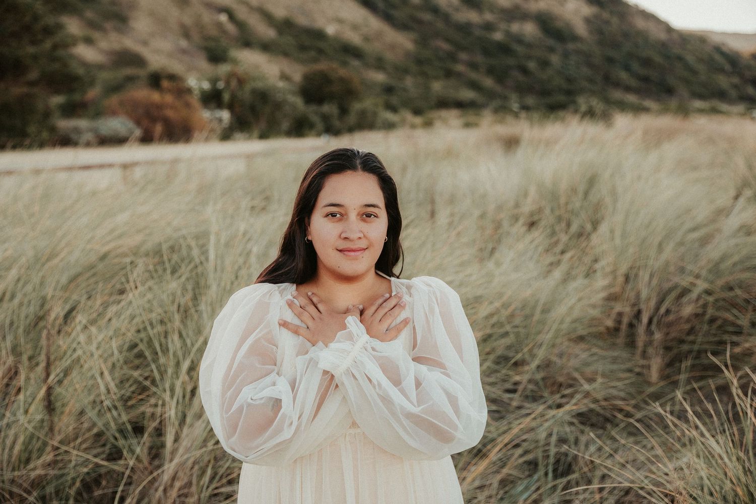 Person in flowing white dress poses with hands crossed over heart outdoors.