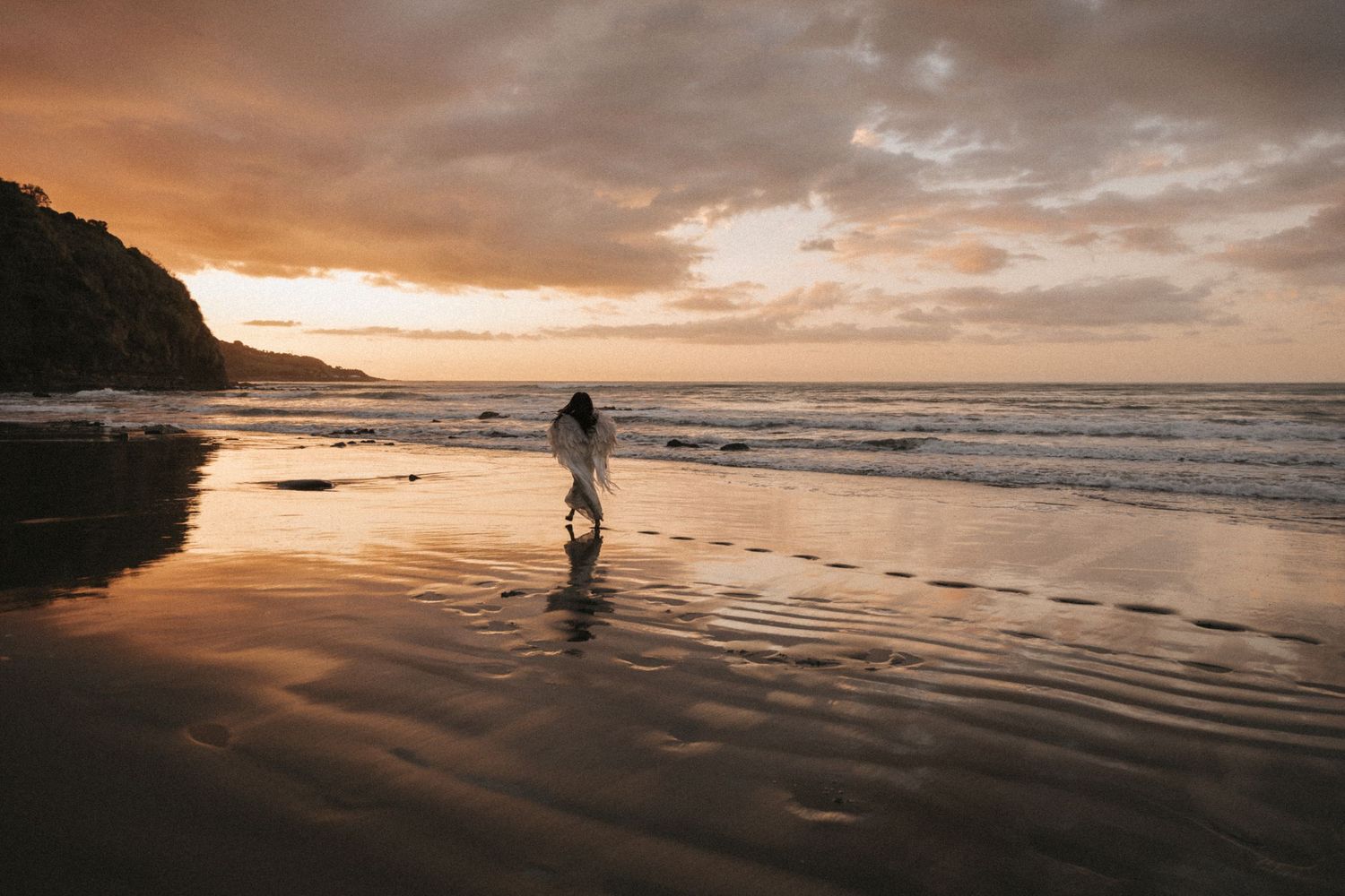 A silhouetted figure walks along a dramatic beach shoreline at sunset with golden clouds reflecting on wet sand.