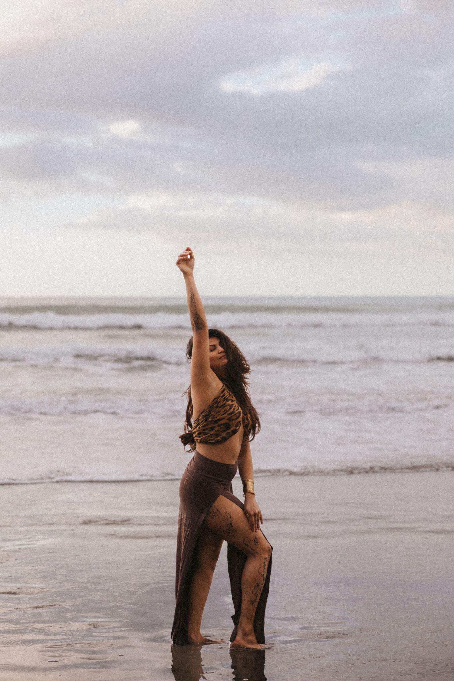 A person in brown clothing dances expressively on a misty beach at sunset, raising their arm in a graceful pose.