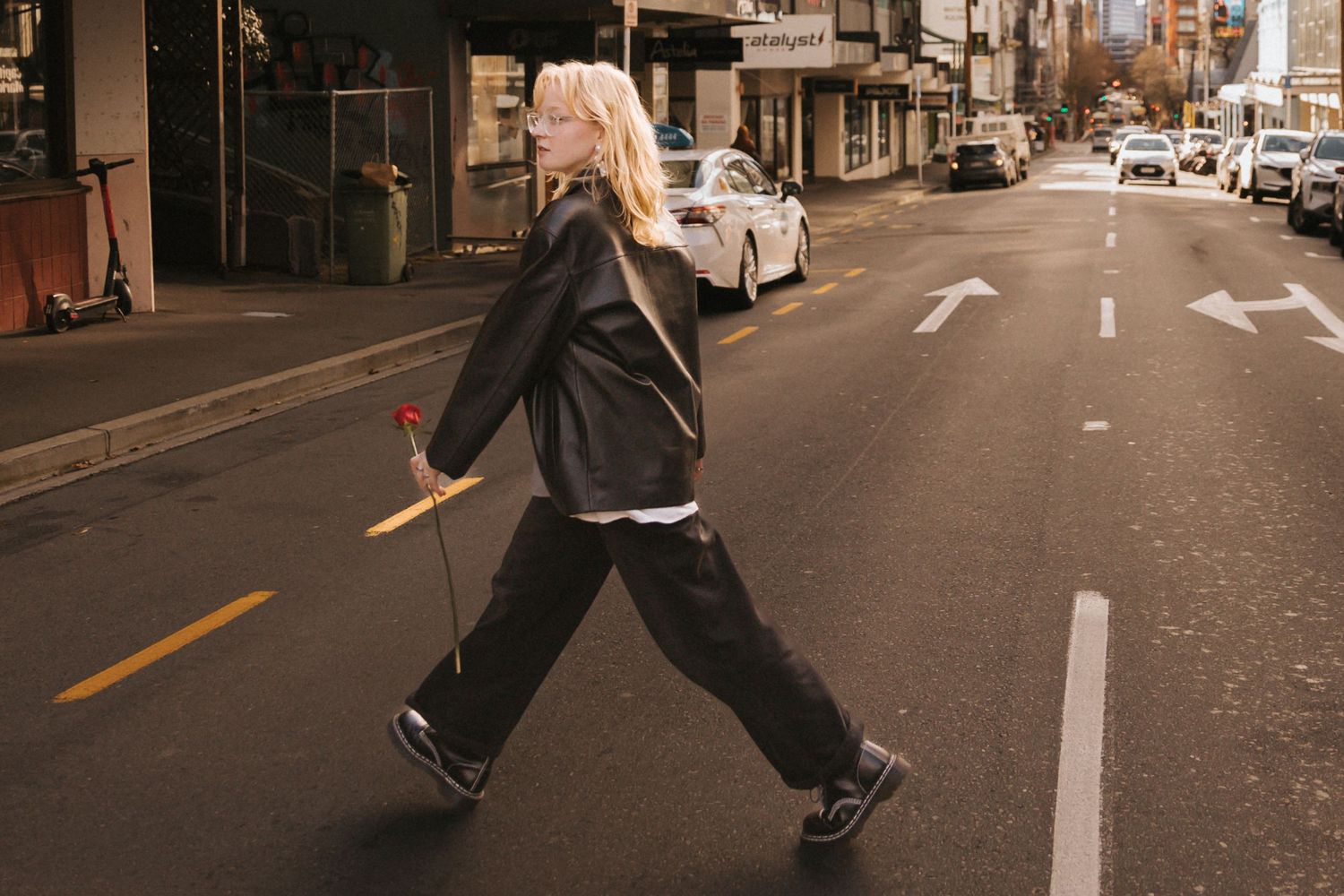 Person wearing dark clothing walks down an urban street with buildings and parked cars visible in the background.