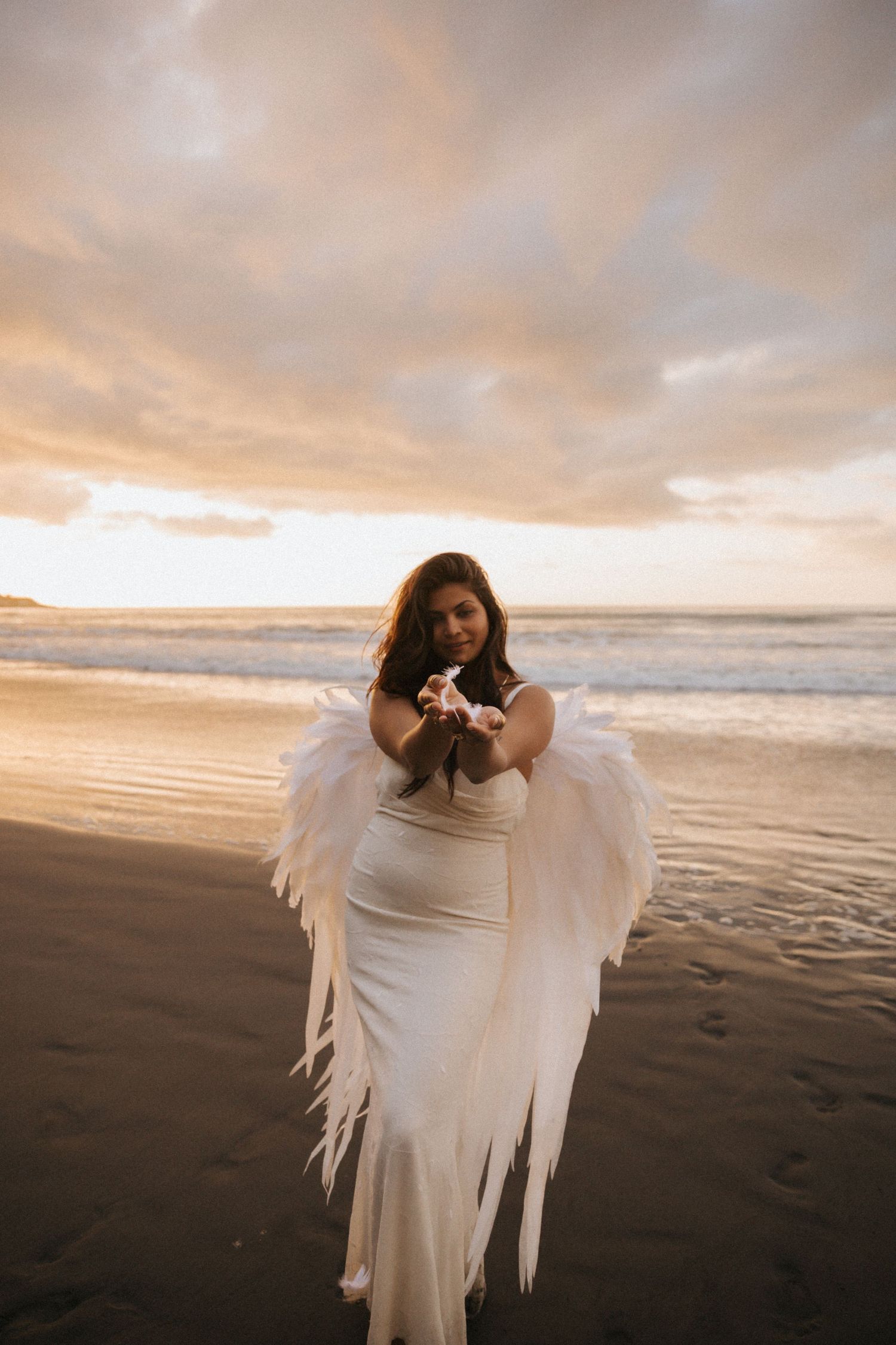 A figure in a flowing white dress and angel wings poses dramatically on a beach at sunset.