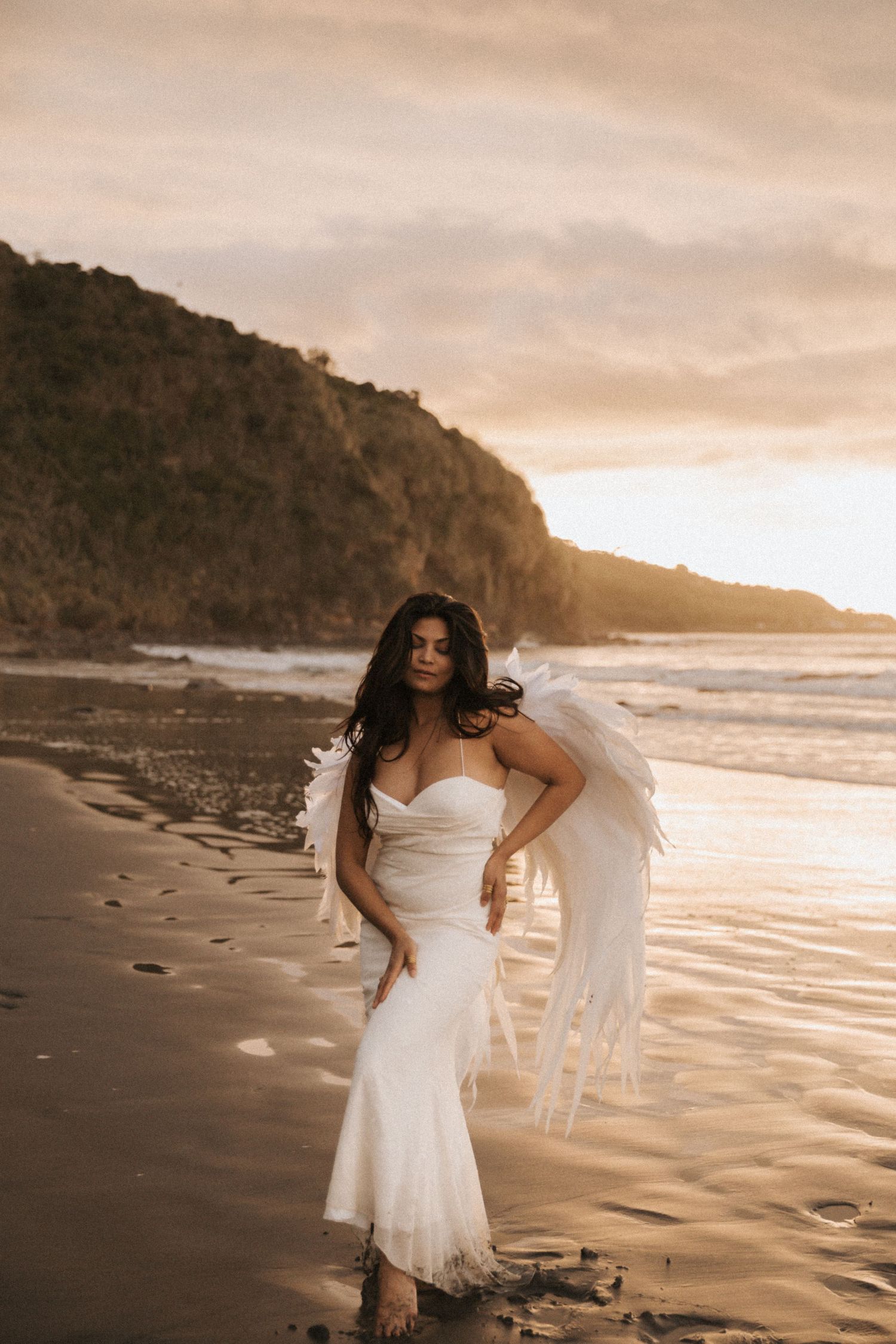 Ethereal beach photoshoot at sunset with flowing white dress against dramatic coastal cliffs and golden sand.