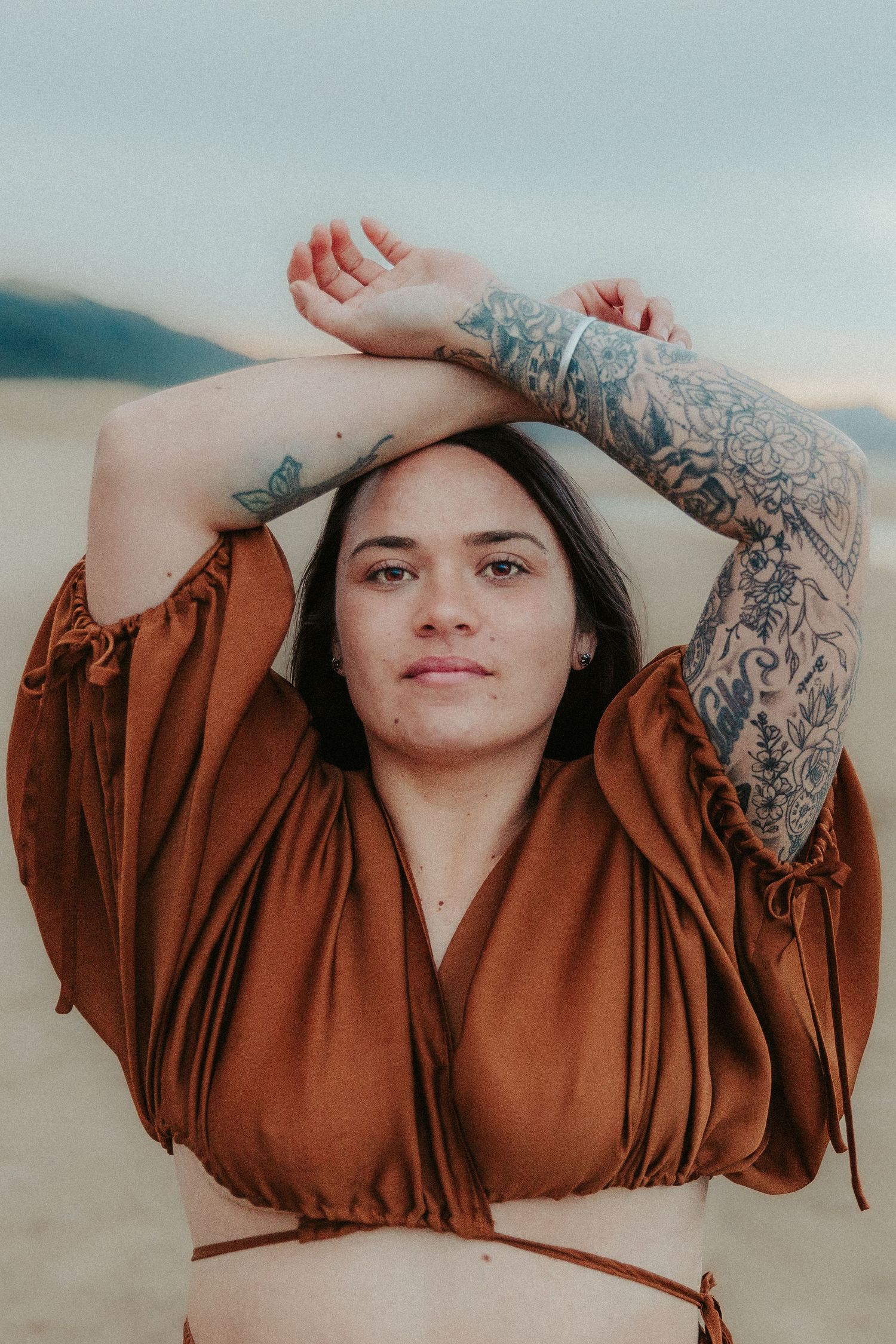 Artistic portrait showcasing intricate tattoo sleeves and rust-colored flowing dress on beach.