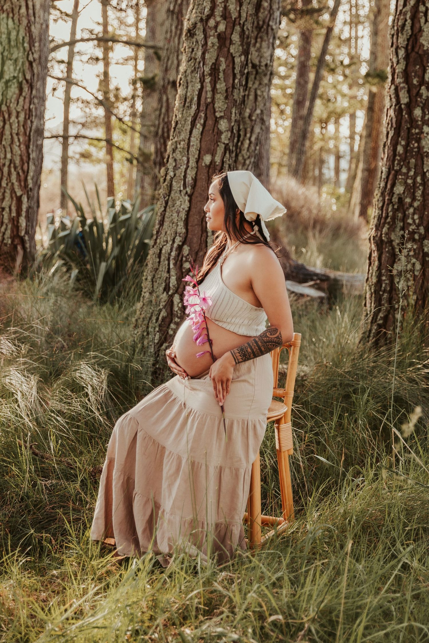 Maternity photo in a forest setting with flowing pink dress and white top against tall pine trees in golden sunlight.