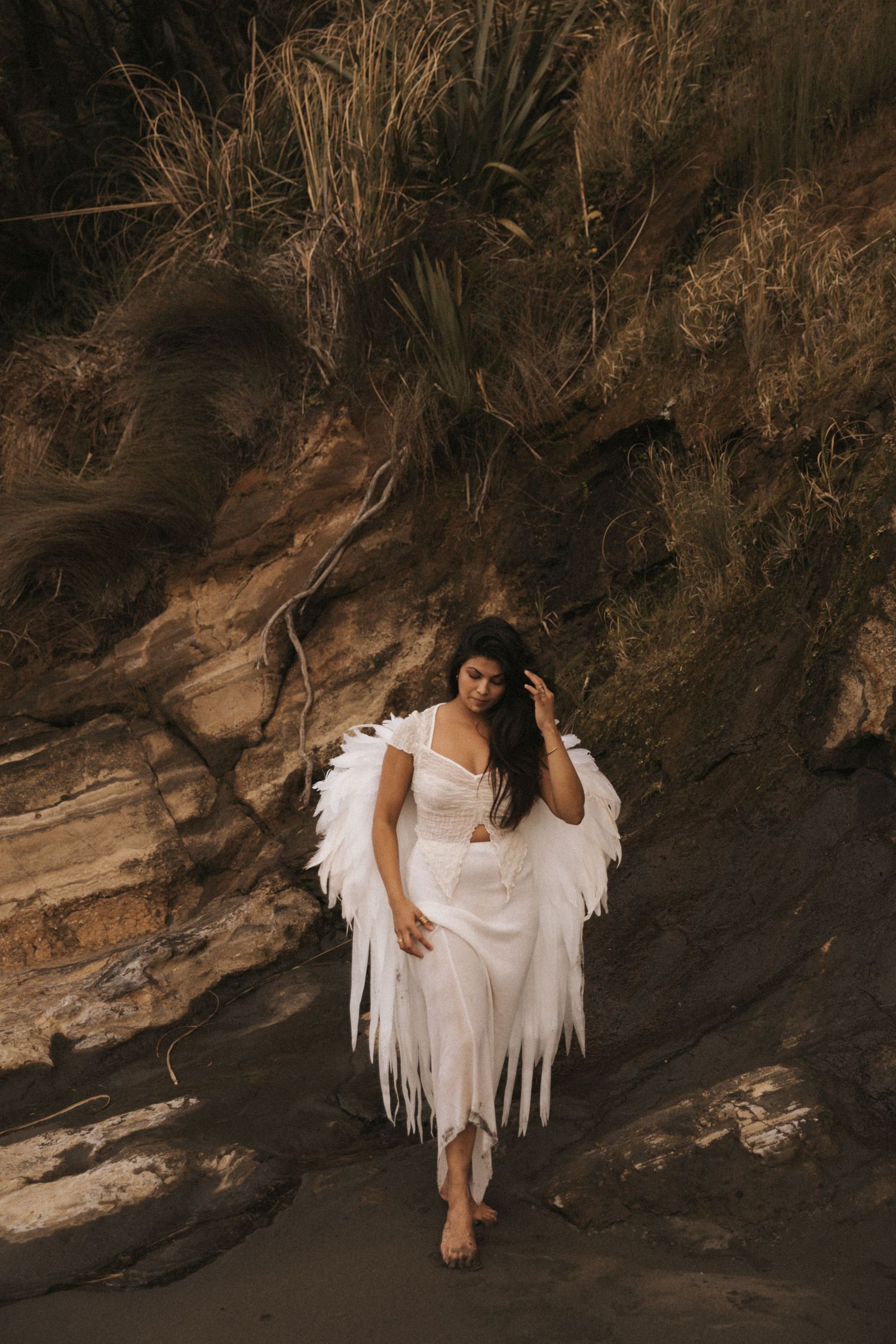 A figure in a flowing white dress and angel wings poses barefoot against dramatic rocky cliffs at sunset.