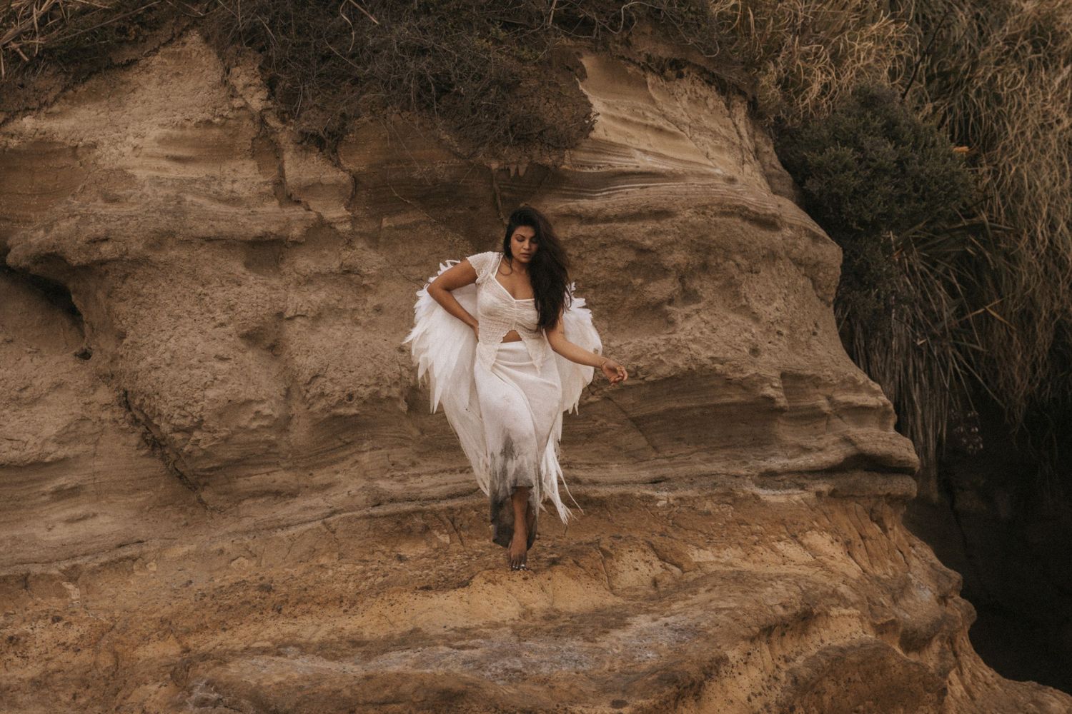 A person in a white dress sits on rocky terrain surrounded by desert landscape during a photoshoot.