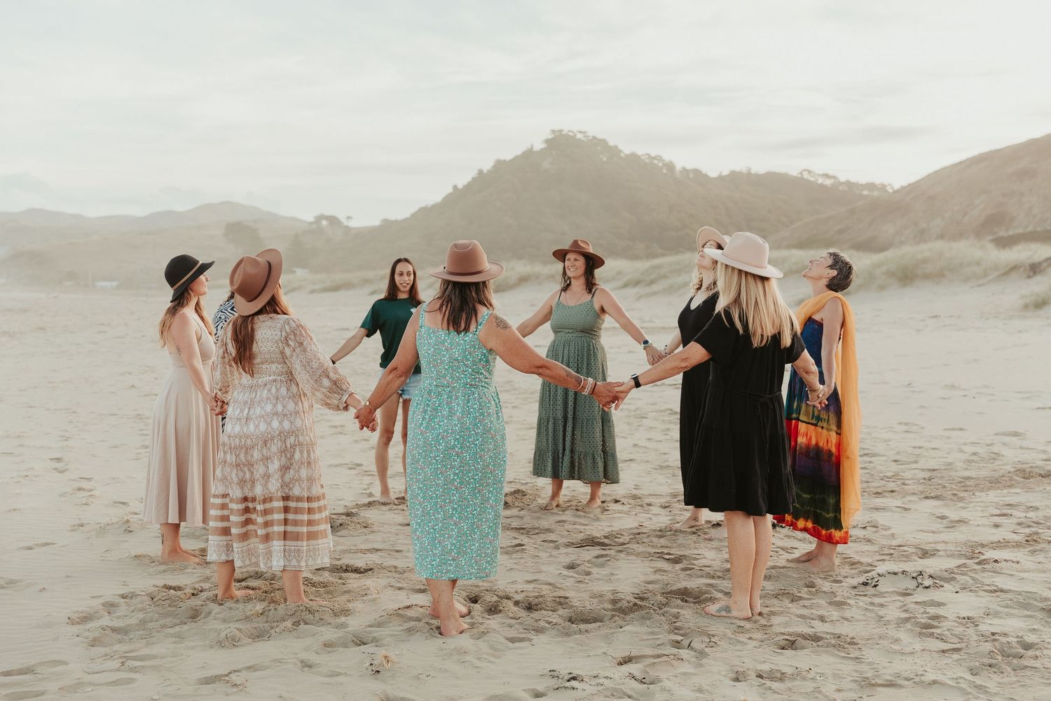 A group of people in flowy dresses dance together on a sandy beach at sunset.