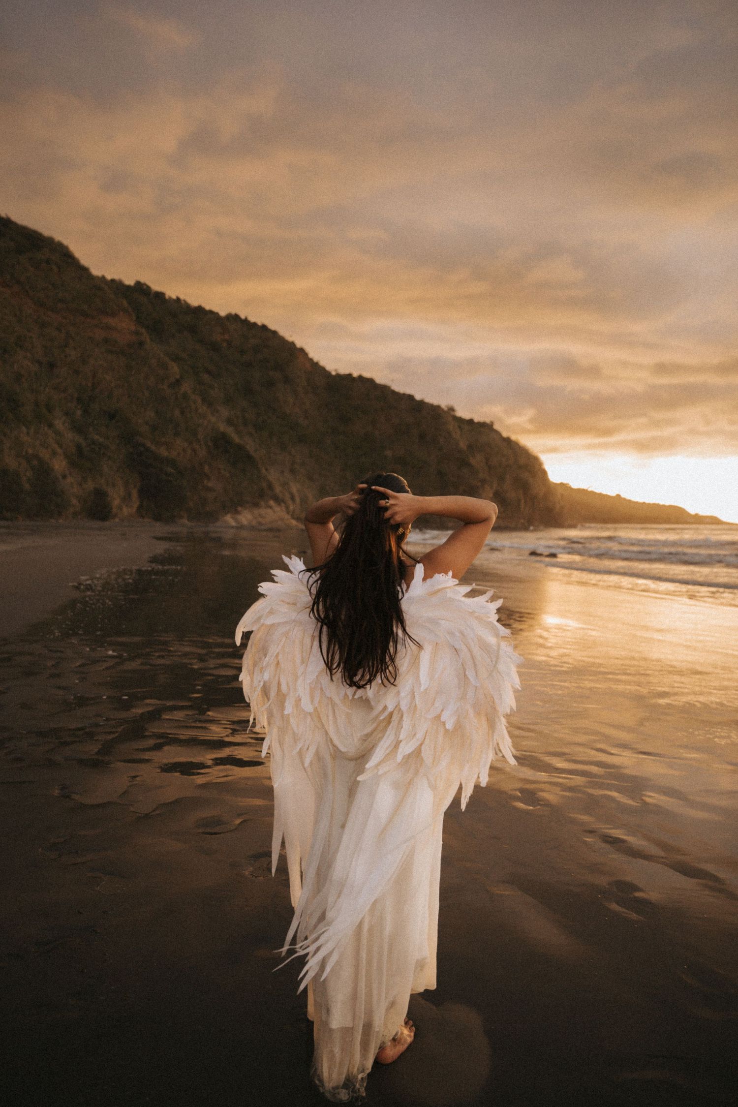 A figure with white angel wings stands on a beach at sunset creating a dreamy silhouette against the golden sky.