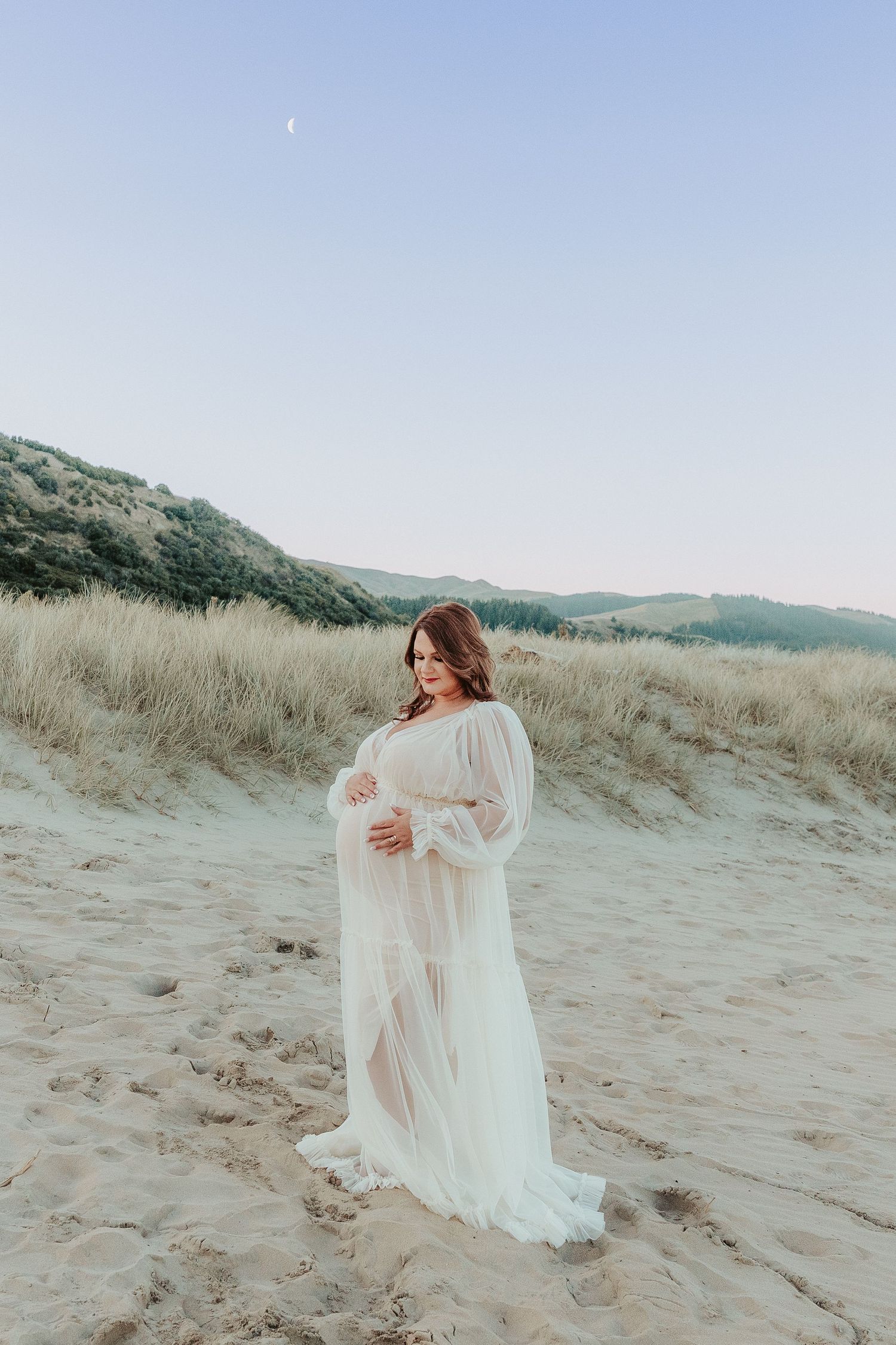 A pregnant woman in a flowing white dress billows in the wind against sandy dunes and distant mountains on a cloudy day.