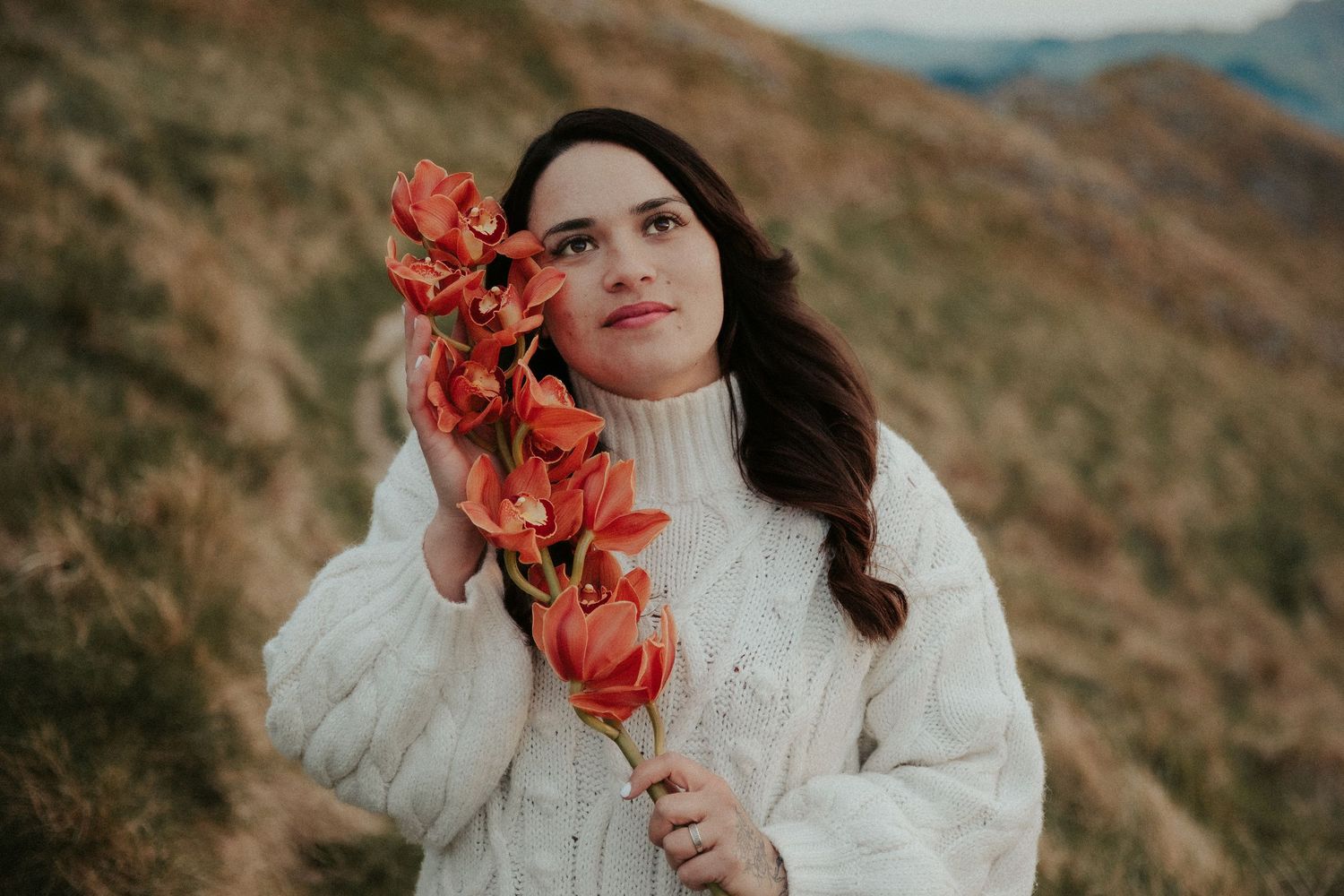 A dreamy portrait in a white sweater holding coral-colored flowers against a mountainous backdrop.