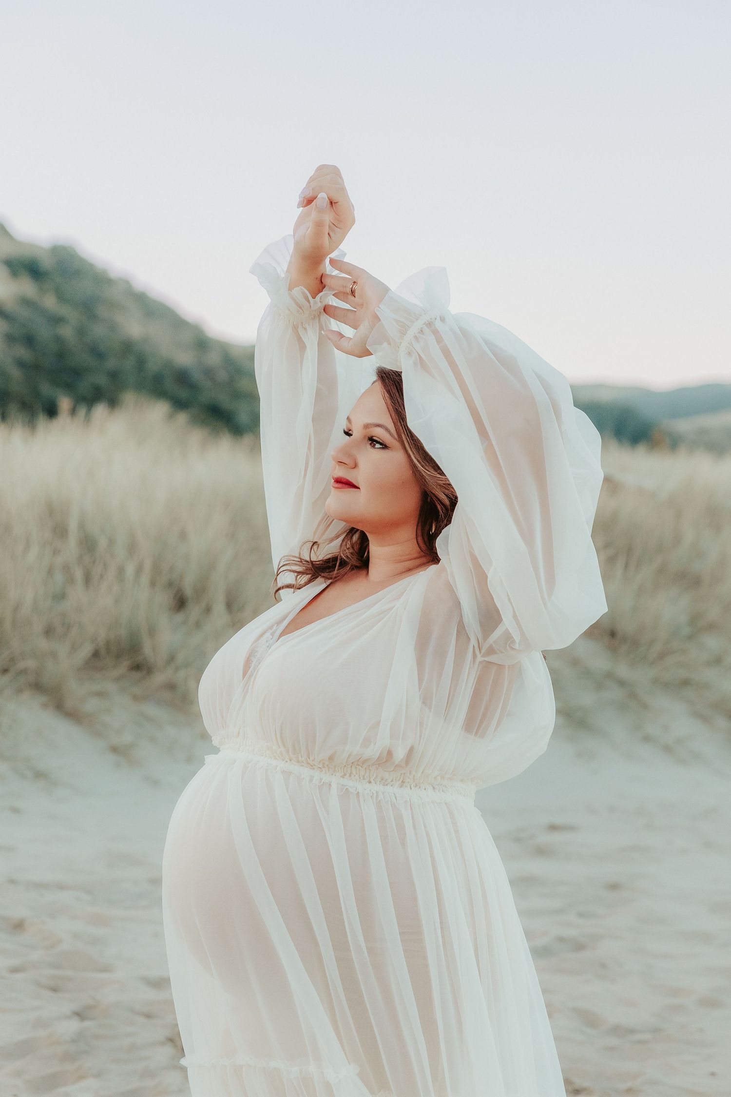 Ethereal maternity photo of woman in flowing white dress posing gracefully in golden field at sunset.