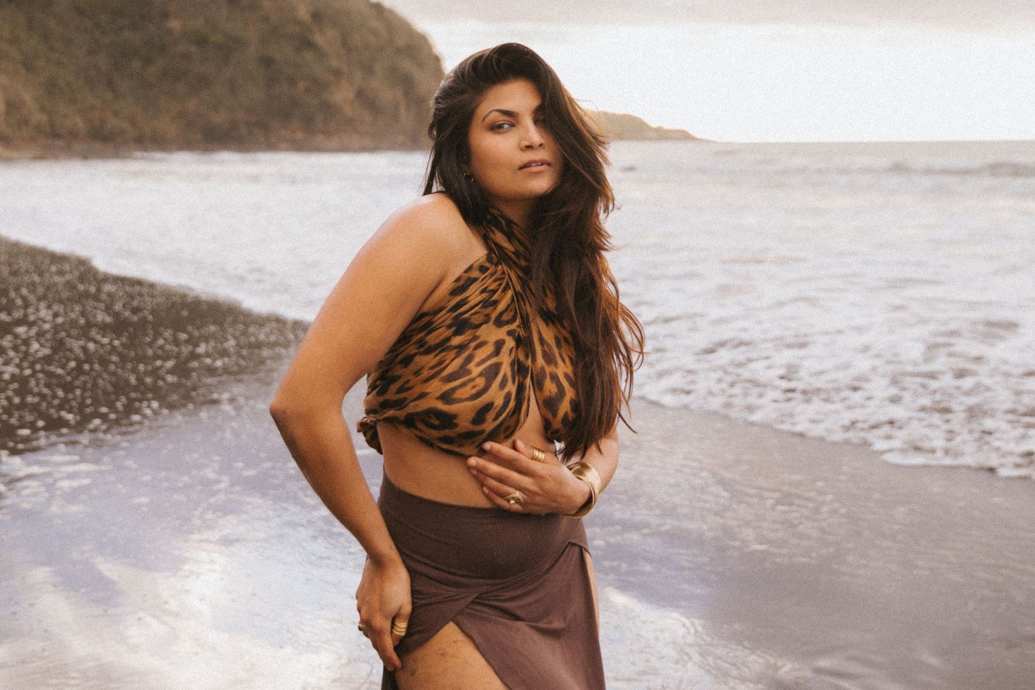 A person in a brown outfit poses confidently along a scenic beach shoreline at sunset.