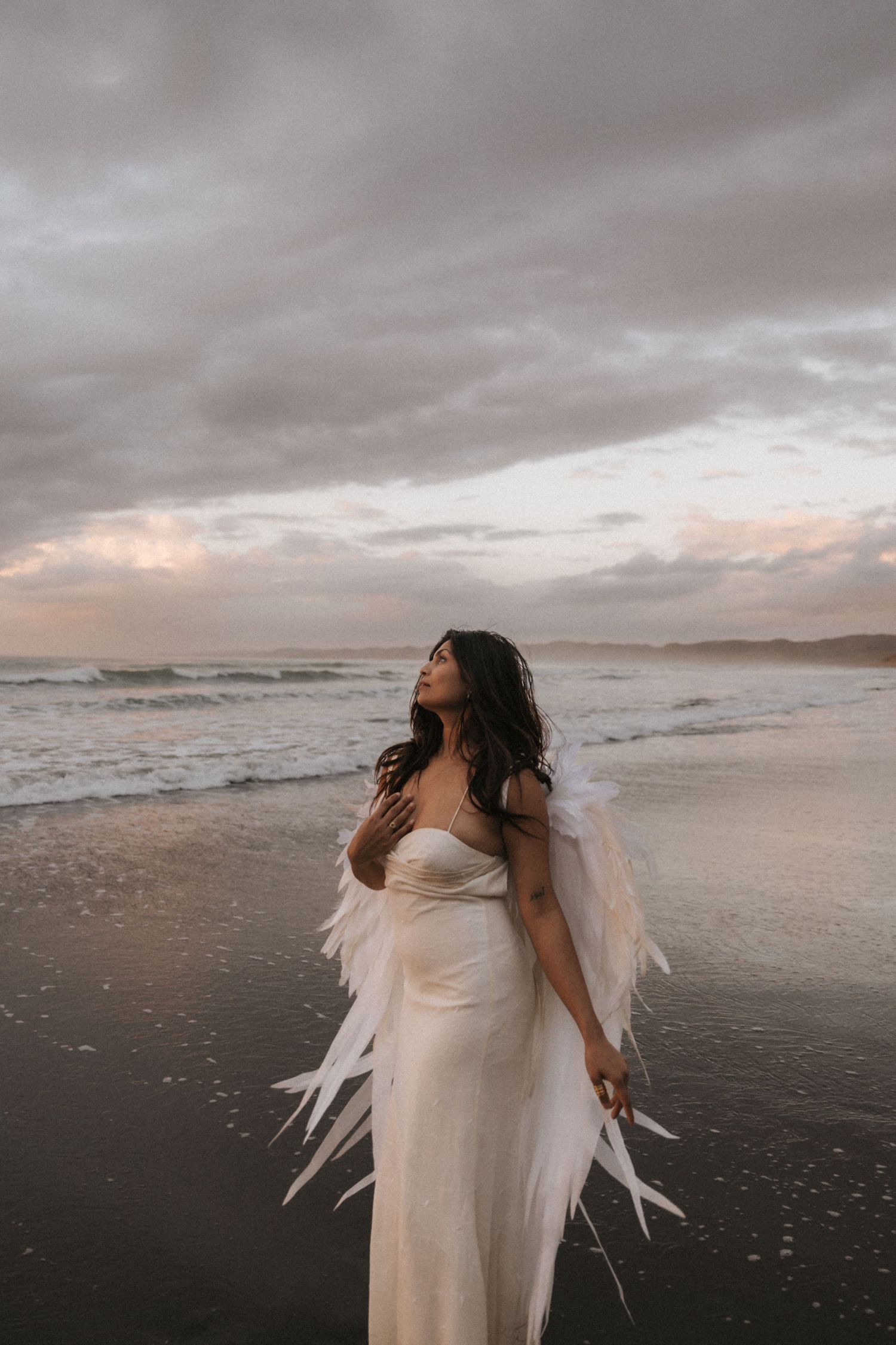 A figure in a flowing white dress with feathered wings stands on a moody beach at sunset.