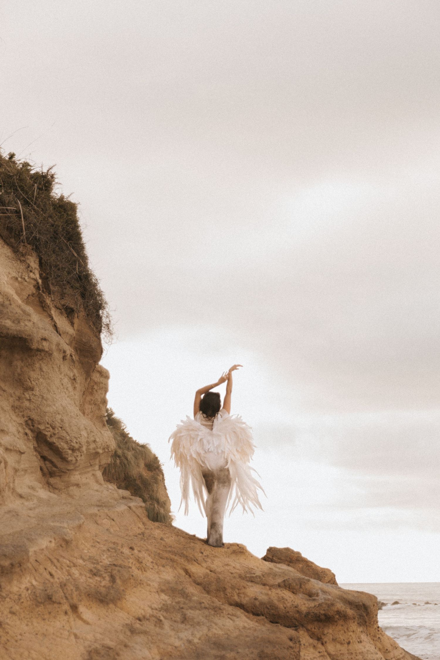 A figure wearing white angel wings stands atop a sandy cliff overlooking the ocean in a moody seascape.