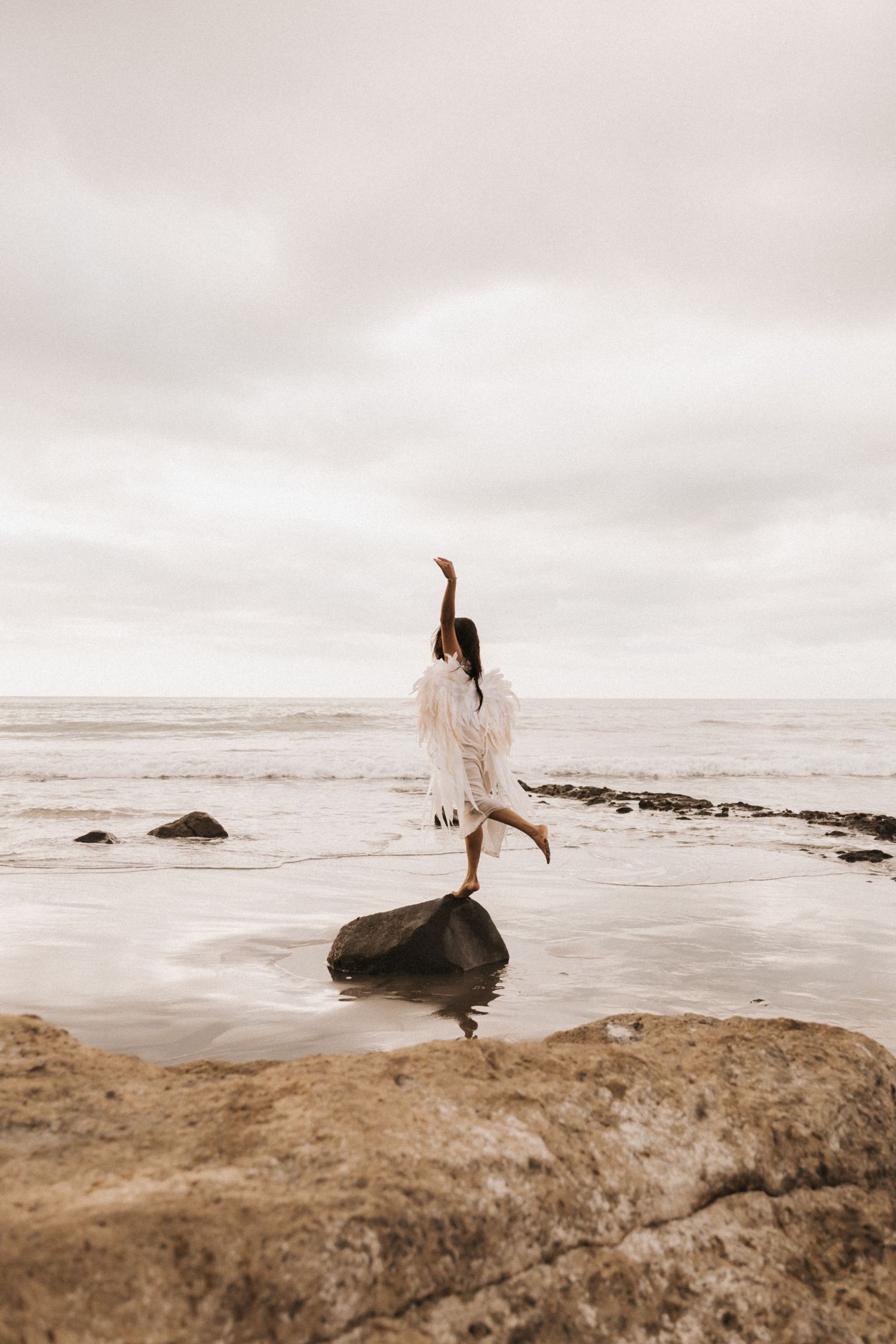 A person in white dress stands on coastal rocks overlooking a misty ocean view at sunset.