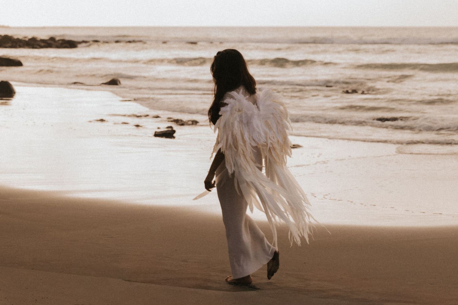 A person in a white dress walks along a sepia-toned beach shoreline at sunset.