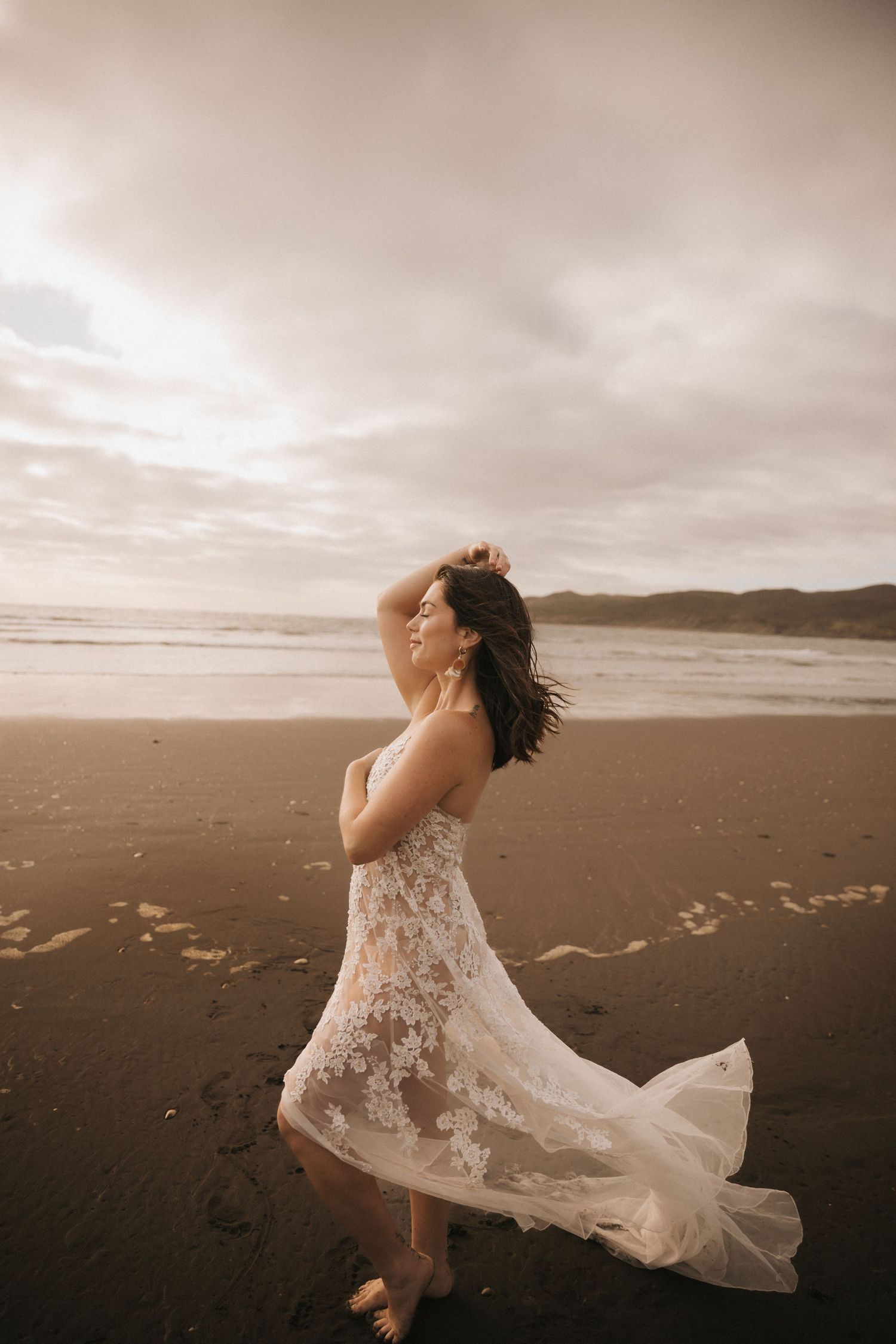 Romantic beach portrait in lace wedding dress with dramatic cloudy sky backdrop.