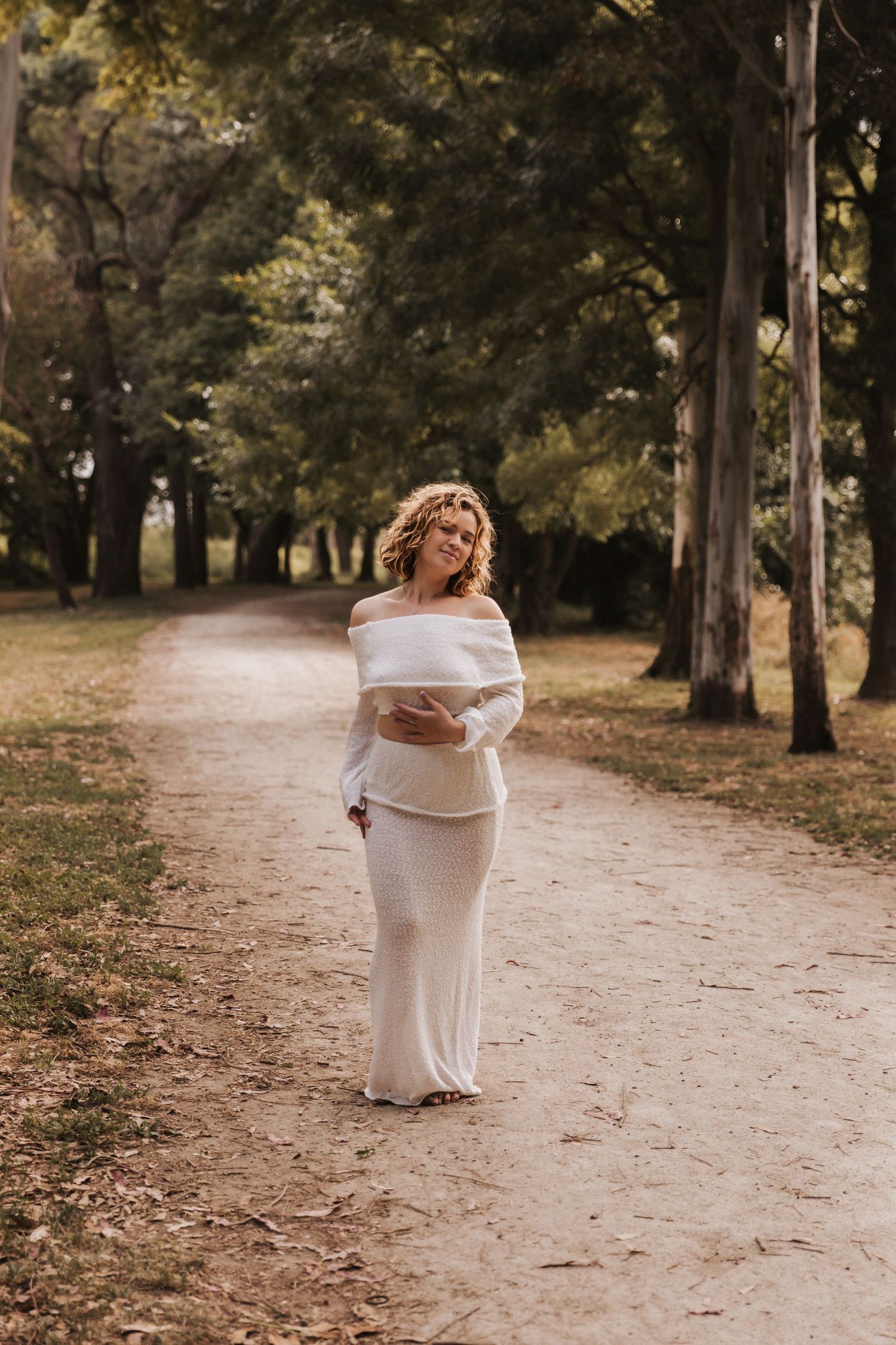Woman in flowing white dress stands on dirt path surrounded by autumn trees.
