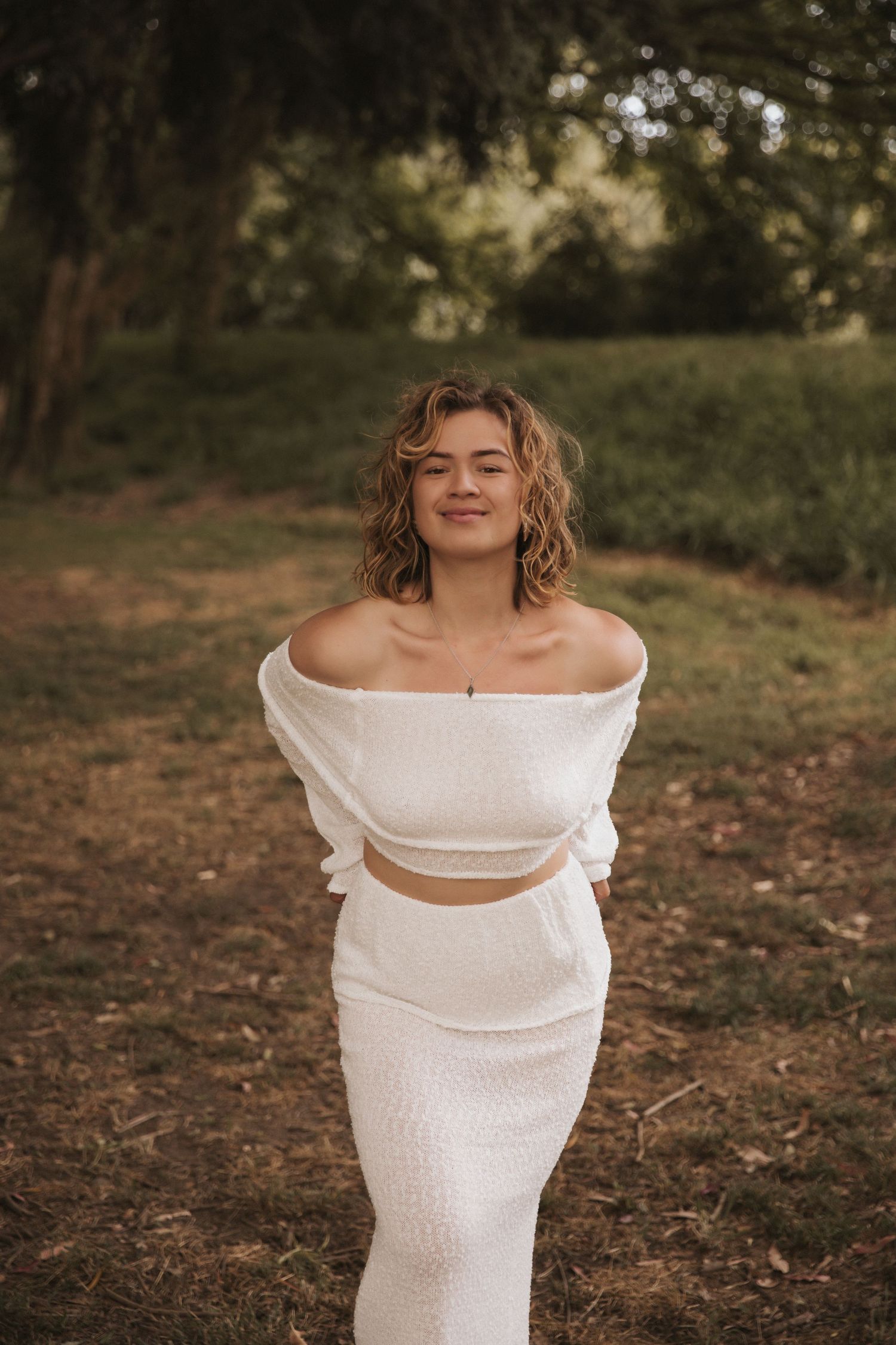 Figure in white two-piece outfit poses against natural outdoor setting.