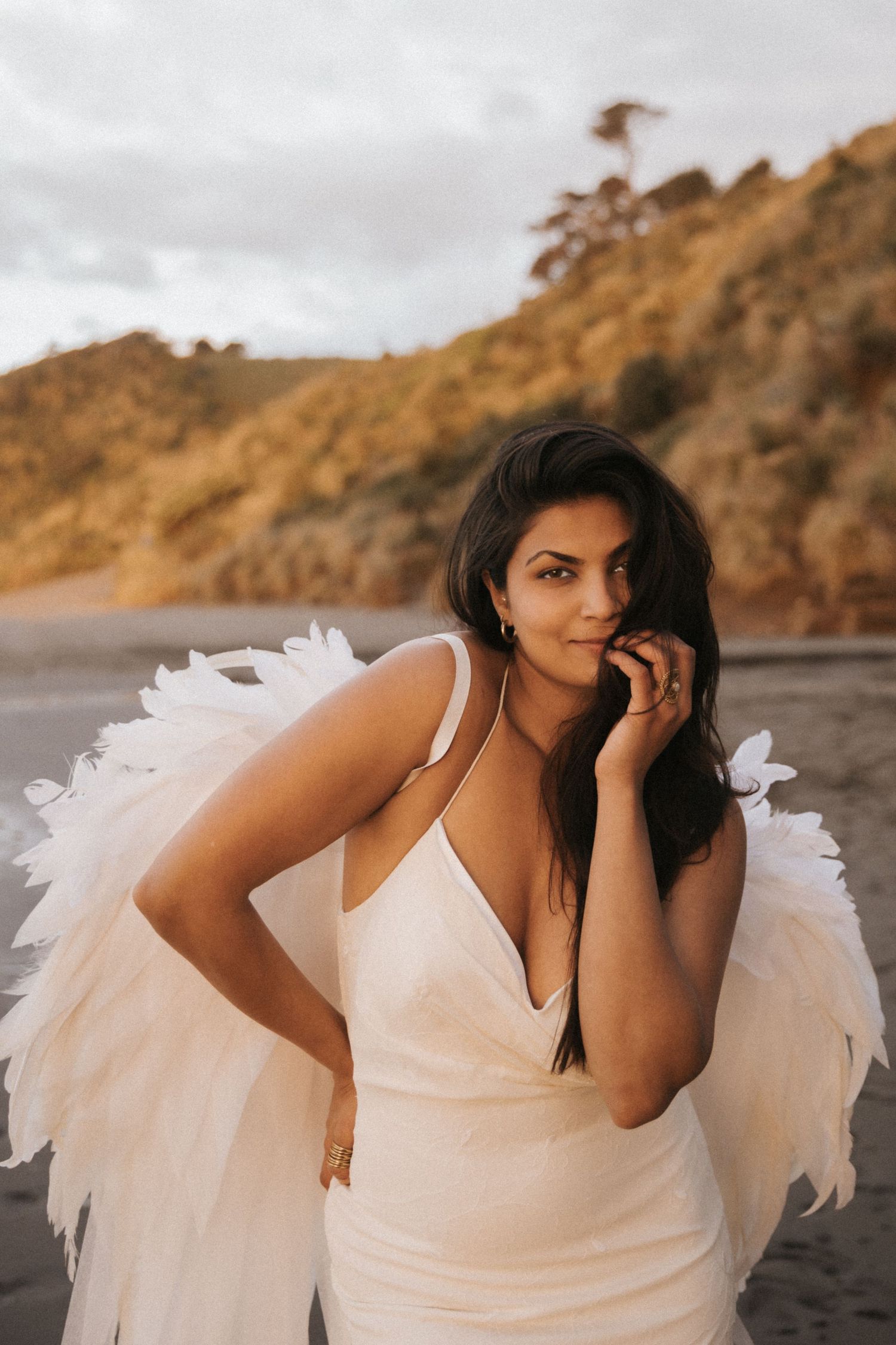 A figure in a white dress with feathered angel wings poses on a dramatic beach at sunset.
