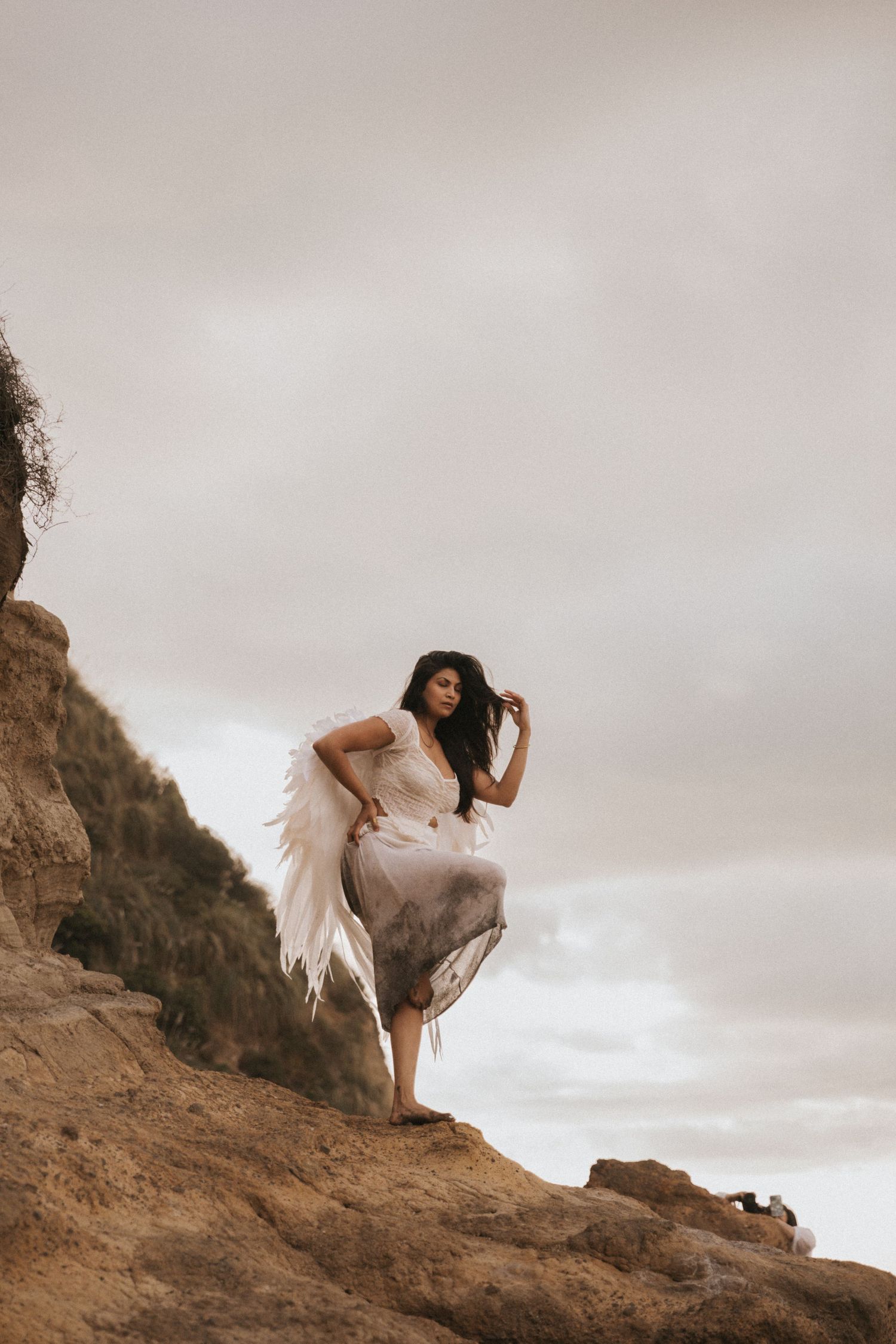 A figure in a flowing white dress poses gracefully on a rocky cliff against a cloudy sky.