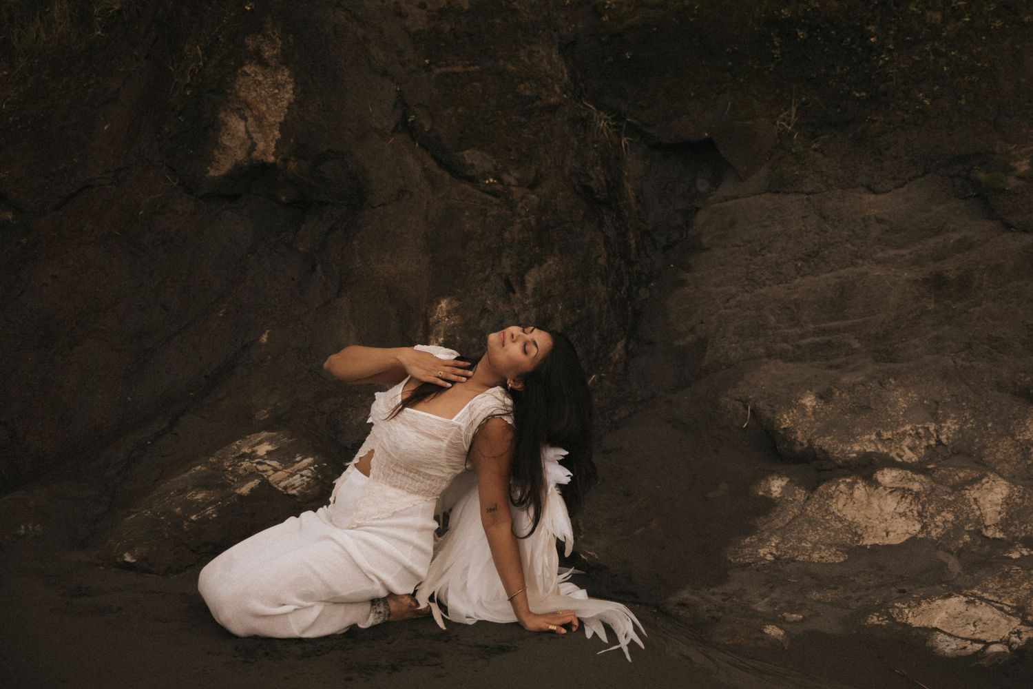 A figure in a flowing white dress poses dramatically against dark rocky cave walls in moody lighting.