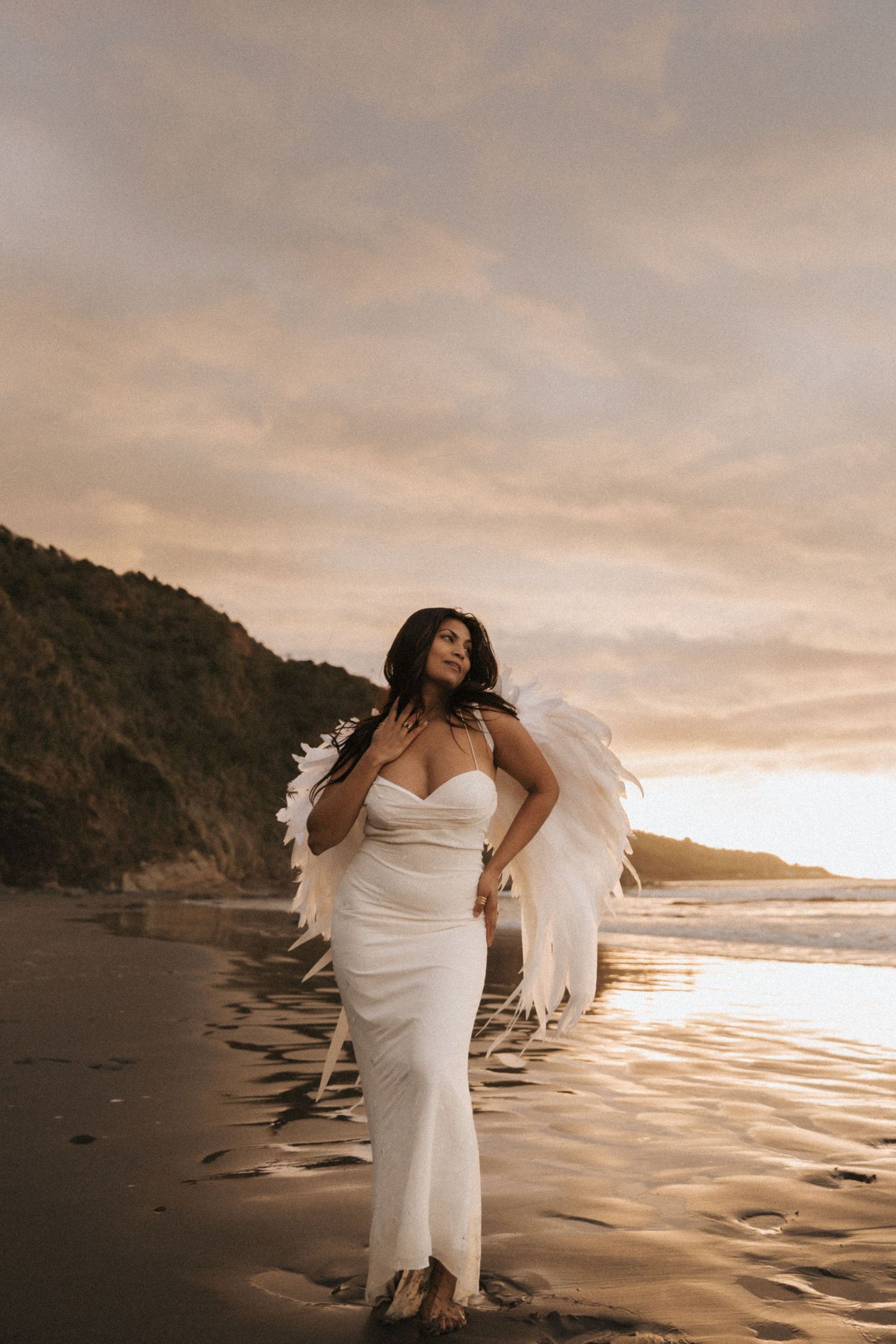 A figure in a flowing white dress poses dramatically on a beach at sunset with angel wings creating an ethereal scene.