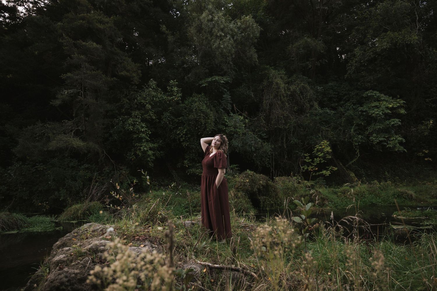An atmospheric photo of a silhouette in a burgundy dress amid wild grasses and dark woods.