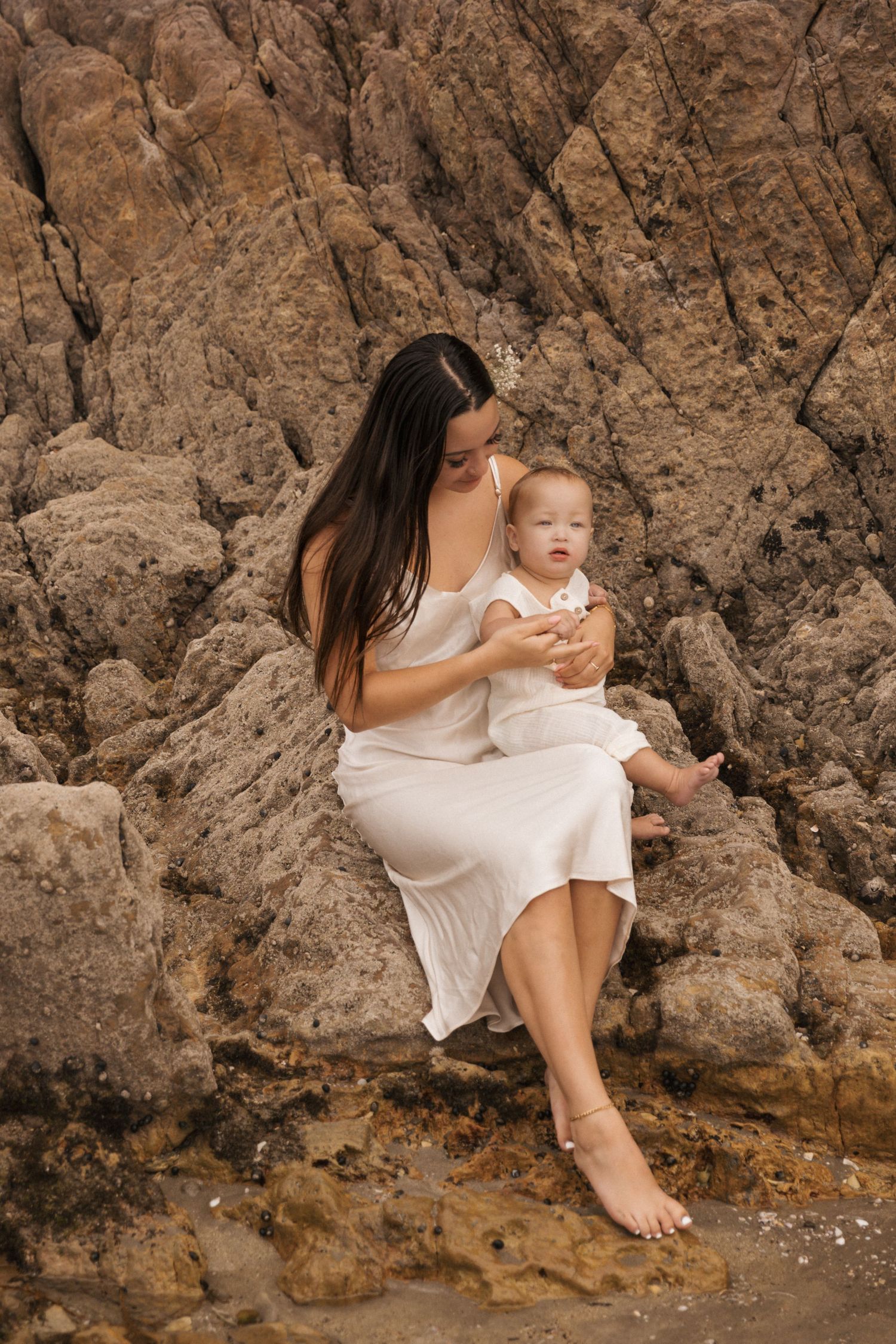 Mother and child in white clothing sitting against rocky cliff backdrop.