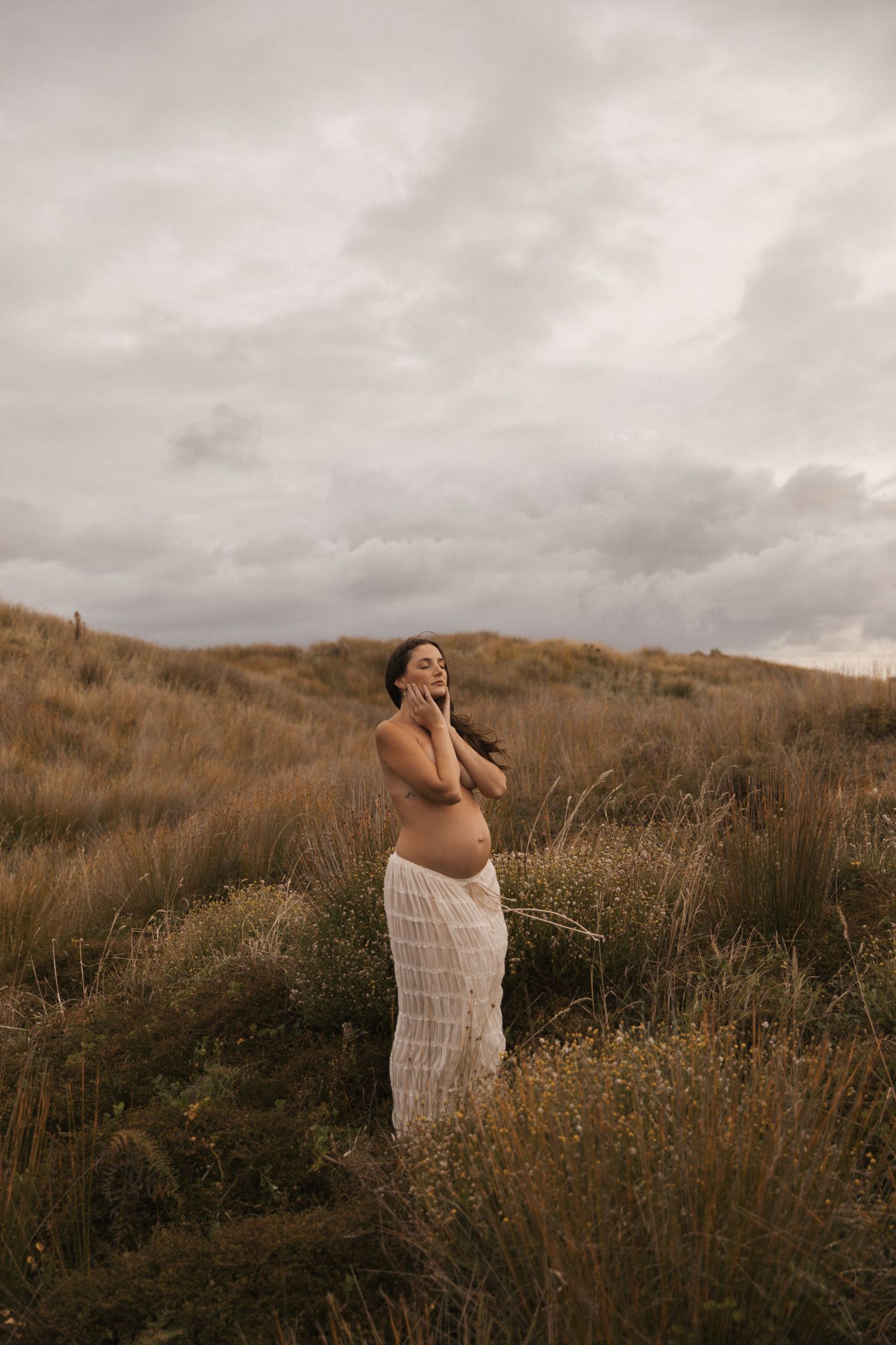Moody maternity photography against cloudy skies and natural terrain.