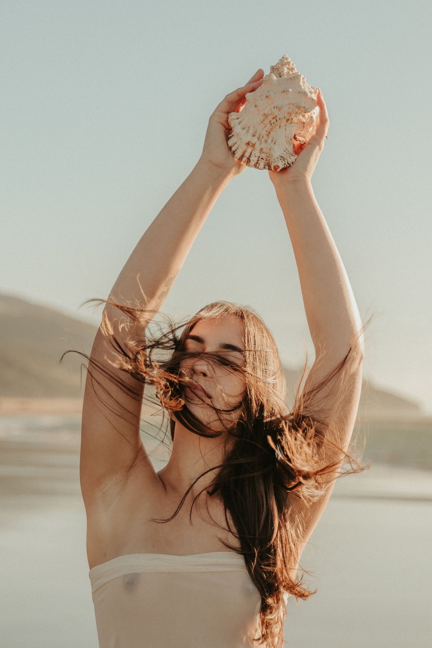 A dreamy beach photoshoot with someone holding a seashell up against the bright sky.