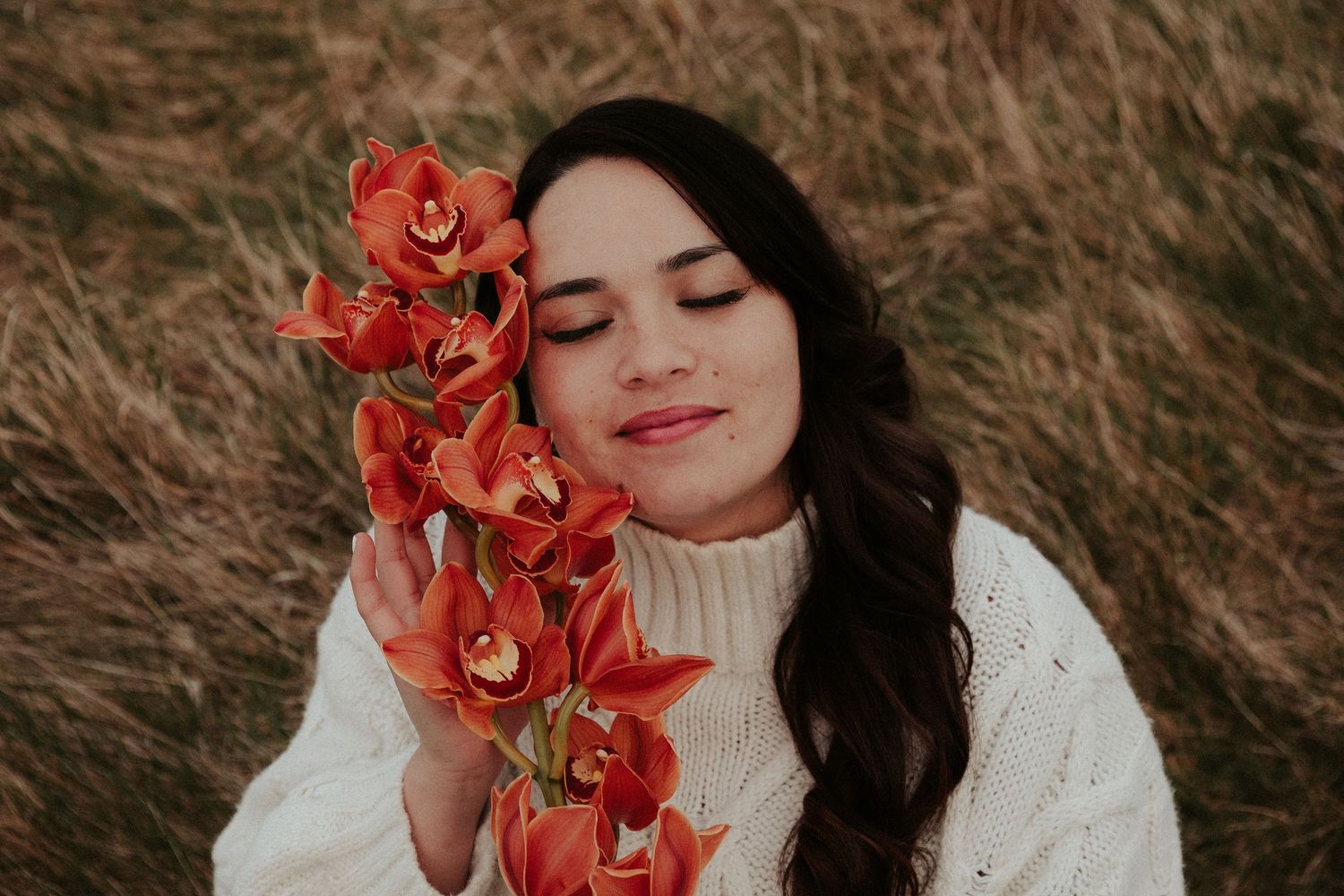 Romantic portrait with coral orchids and white sweater against natural backdrop.