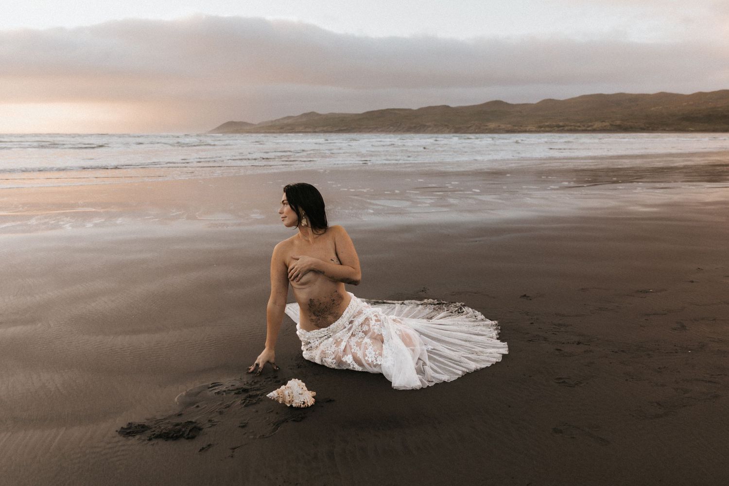 Ethereal beach photography with soft sunset tones and a flowing white dress against coastal mountains.