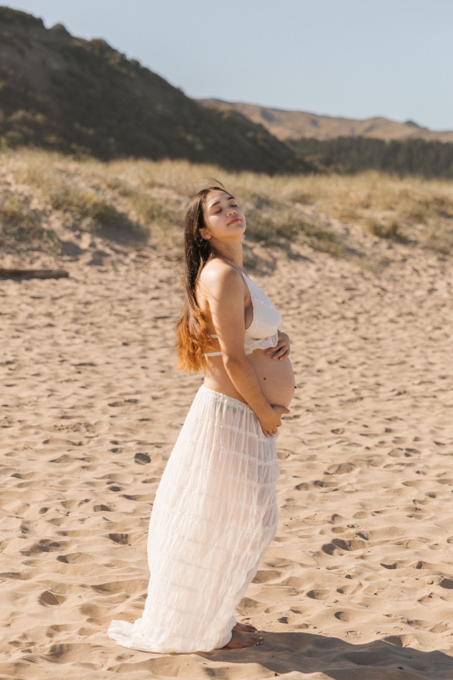 Maternity beach photoshoot with flowing white dress against sandy dunes.