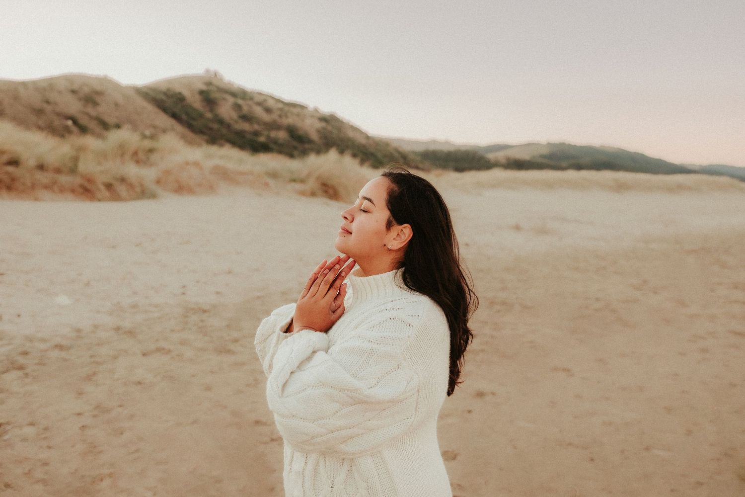 Figure in white sweater moves expressively on beach with mountains in background at sunset.