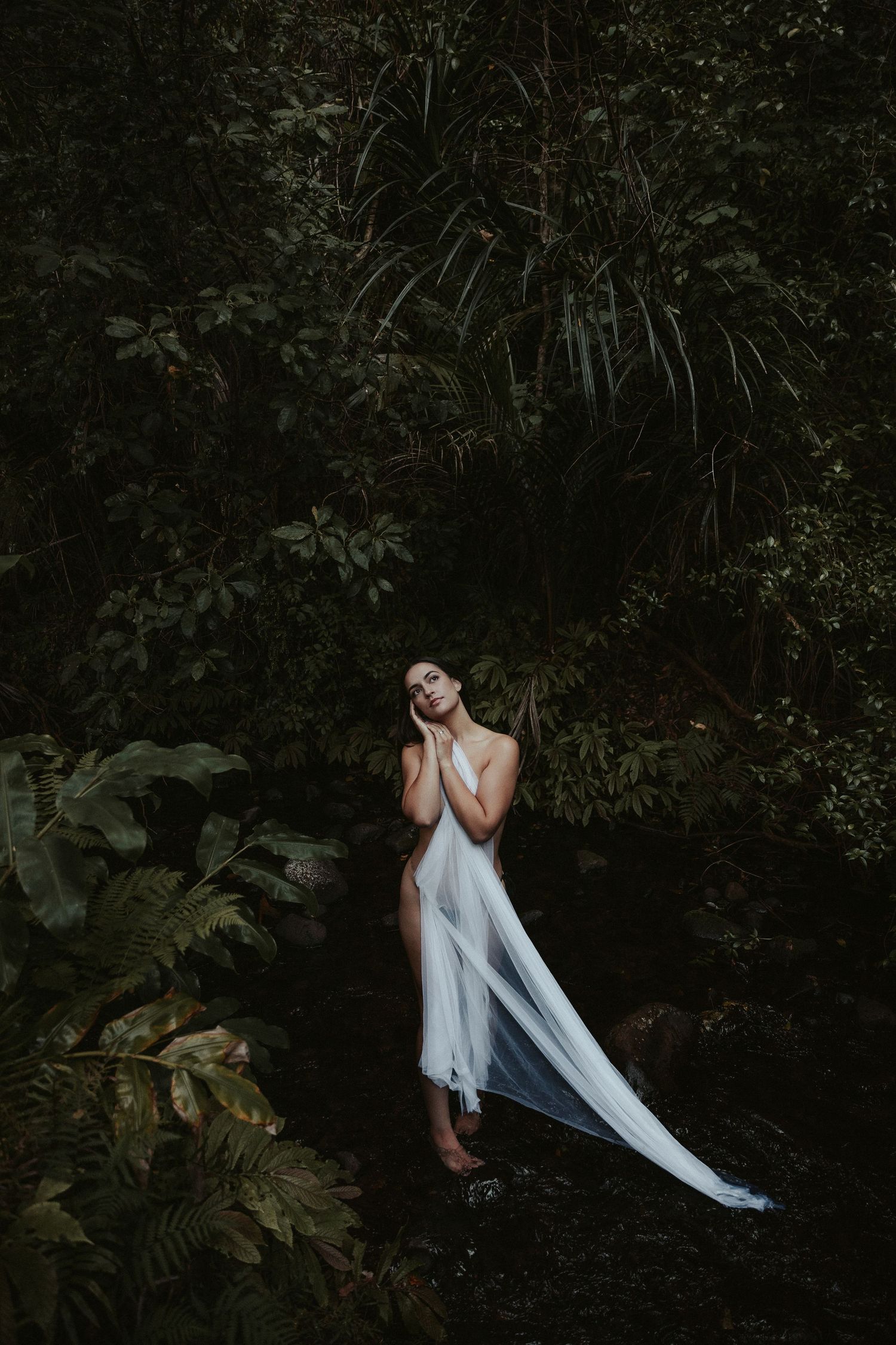 Artistic portrait in dark foliage with flowing white fabric creating ethereal movement against shadowy background.