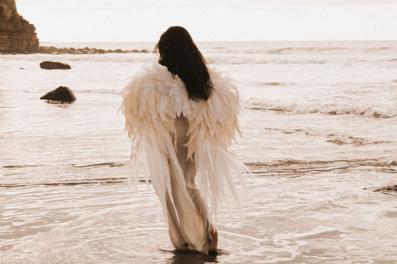 A figure wearing a white dress and angel wings walks along a misty beach at sunset in a dreamy sepia tone.