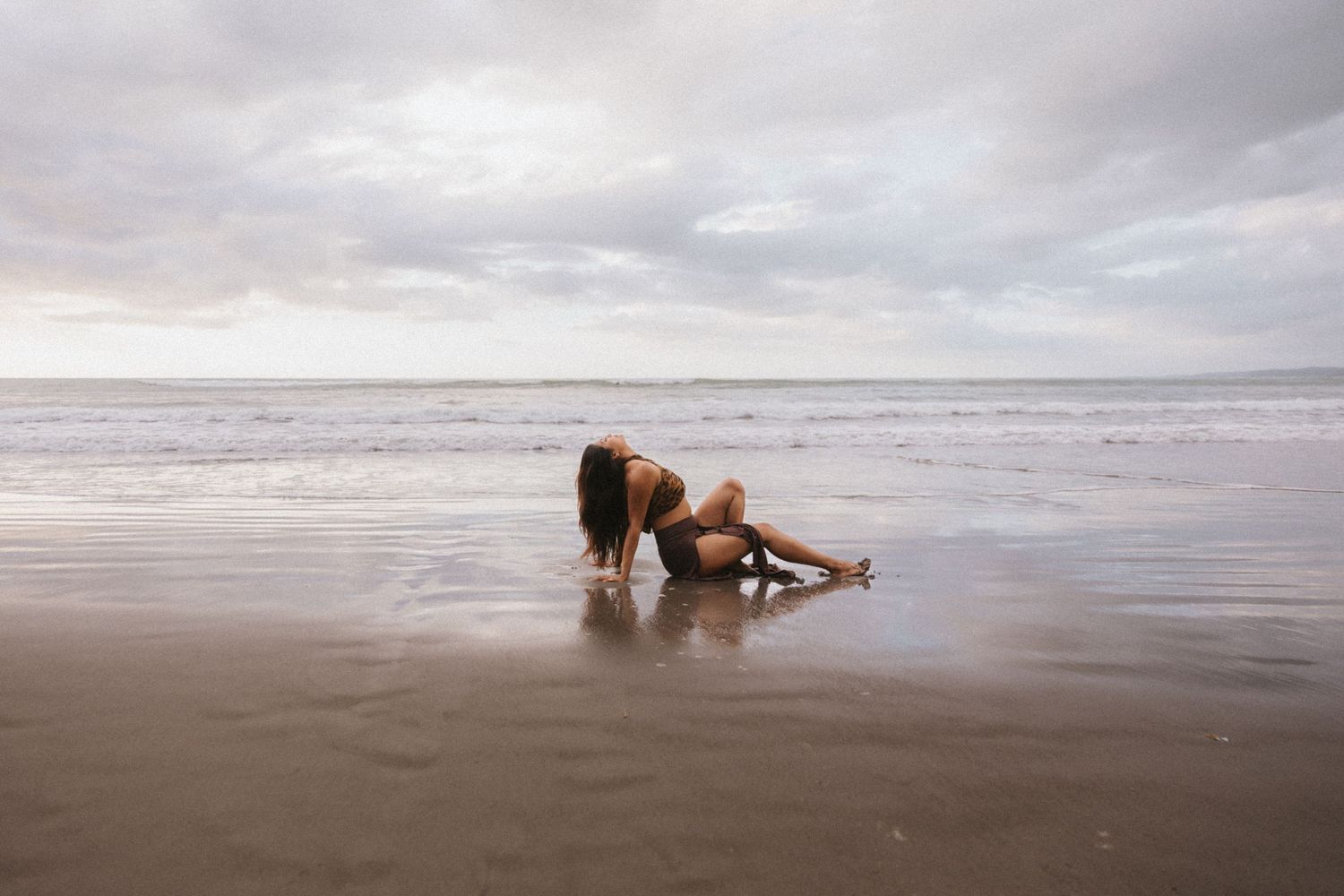 A brown dog plays and splashes in shallow ocean water on an overcast beach day.