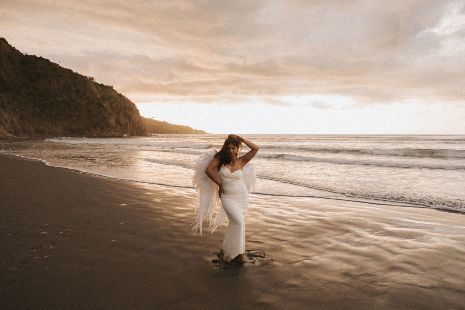 A couple in white wedding attire embraces on a scenic beach at sunset with waves rolling onto the shore.