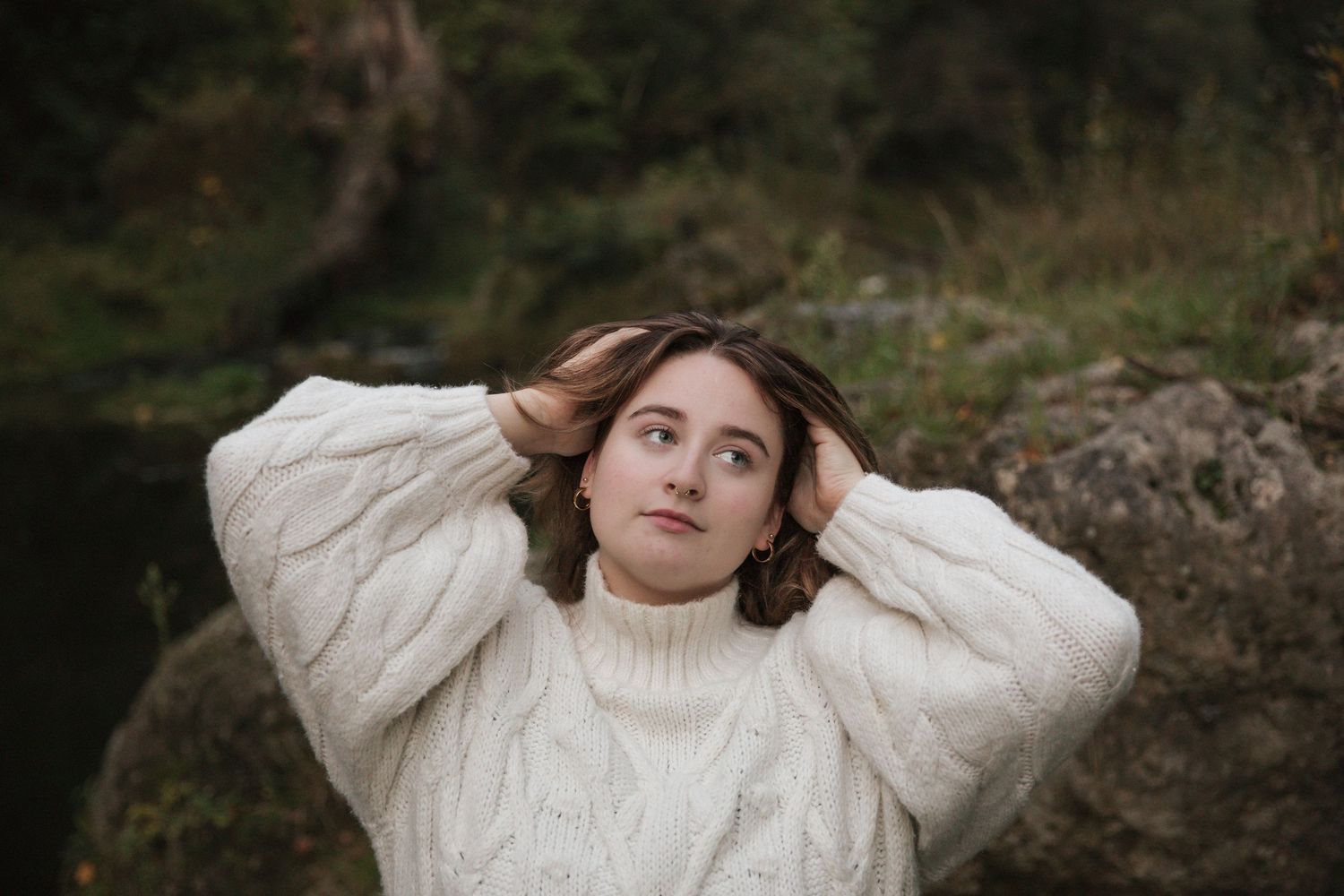 Woman with amazing eyes in cozy cream cable knit sweater poses outdoors against natural backdrop in moody autumn setting.