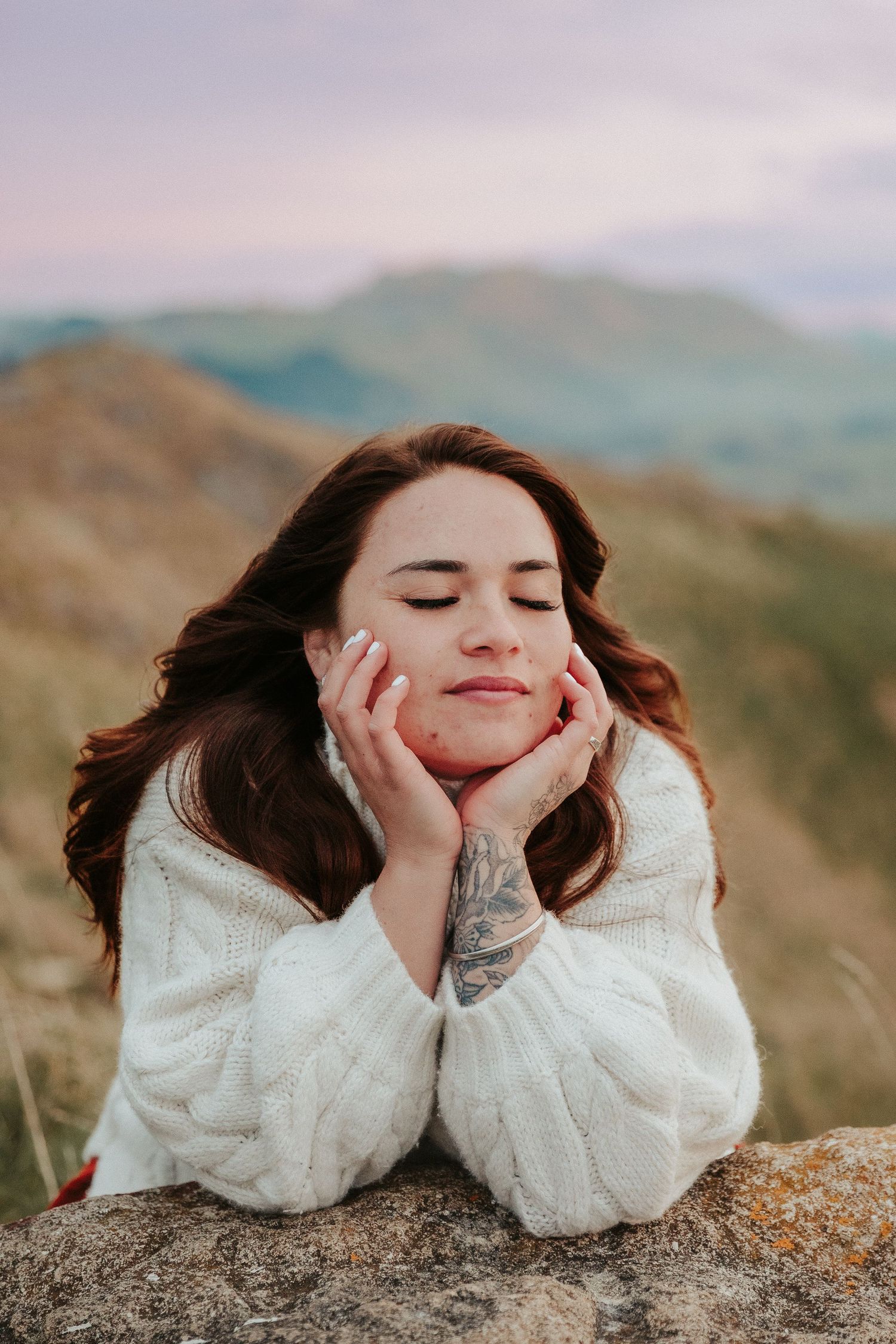 Person in white sweater resting head on hands while lying on rocky surface with mountains in background.